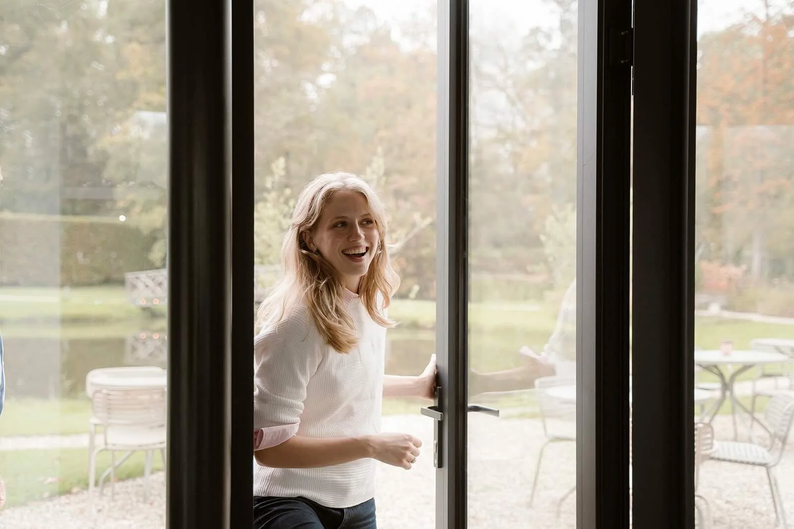 Een lachende vrouw opent een glazen terrasdeur waar zo een workshop plaats zal vinden over persoonlijk leiderschap, buiten is een tuin met tuinmeubelen en bomen te zien.