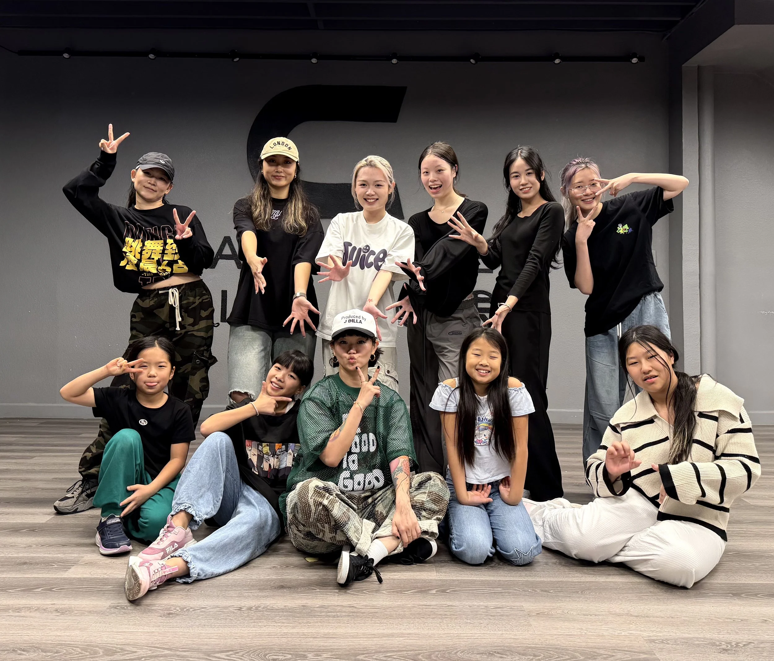 Group of young girls posing together in a dance studio with a gray wall and wooden floor, some standing and some sitting on the floor, smiling and making poses.
