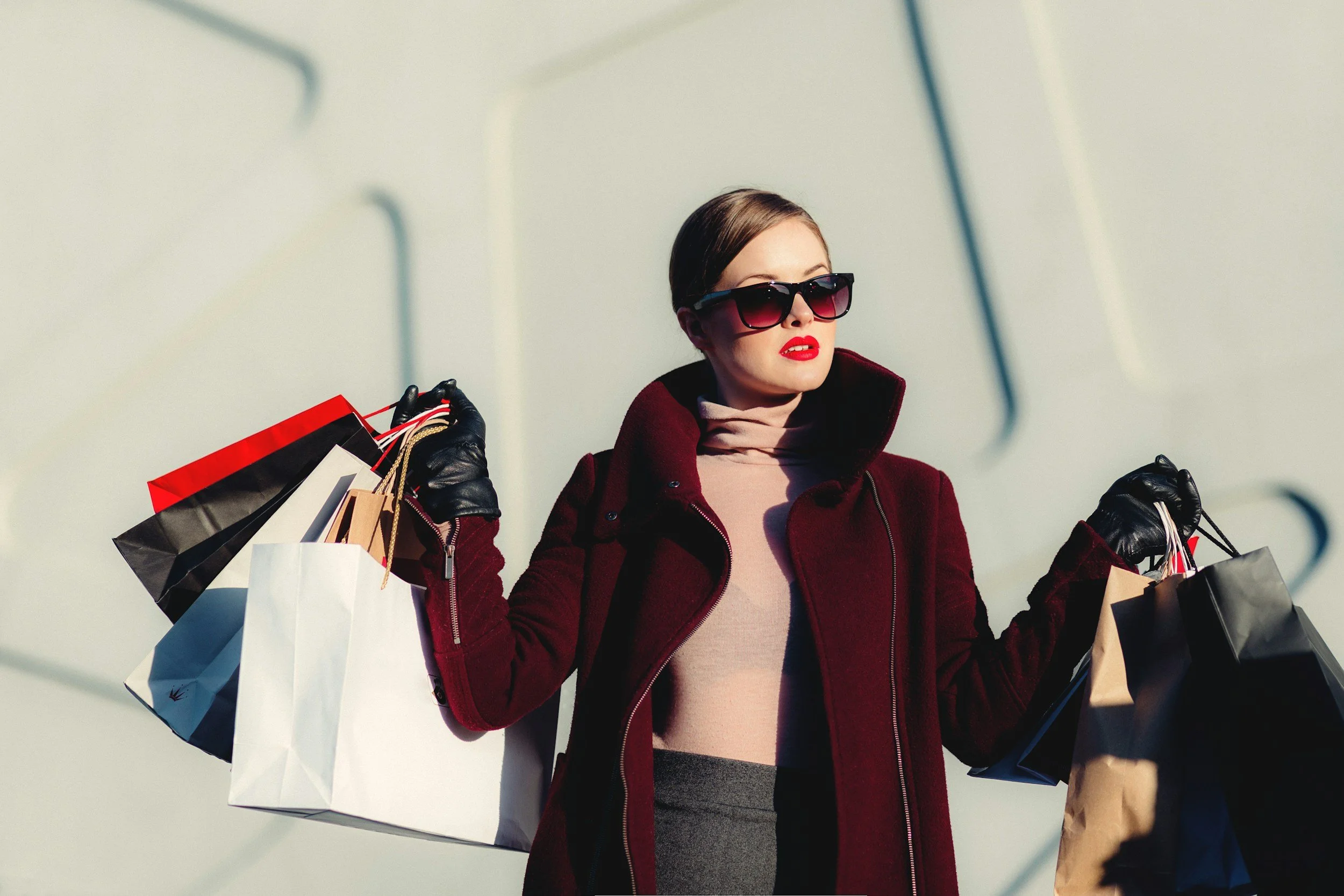 Woman in sunglasses and a red coat holding multiple shopping bags outdoors.