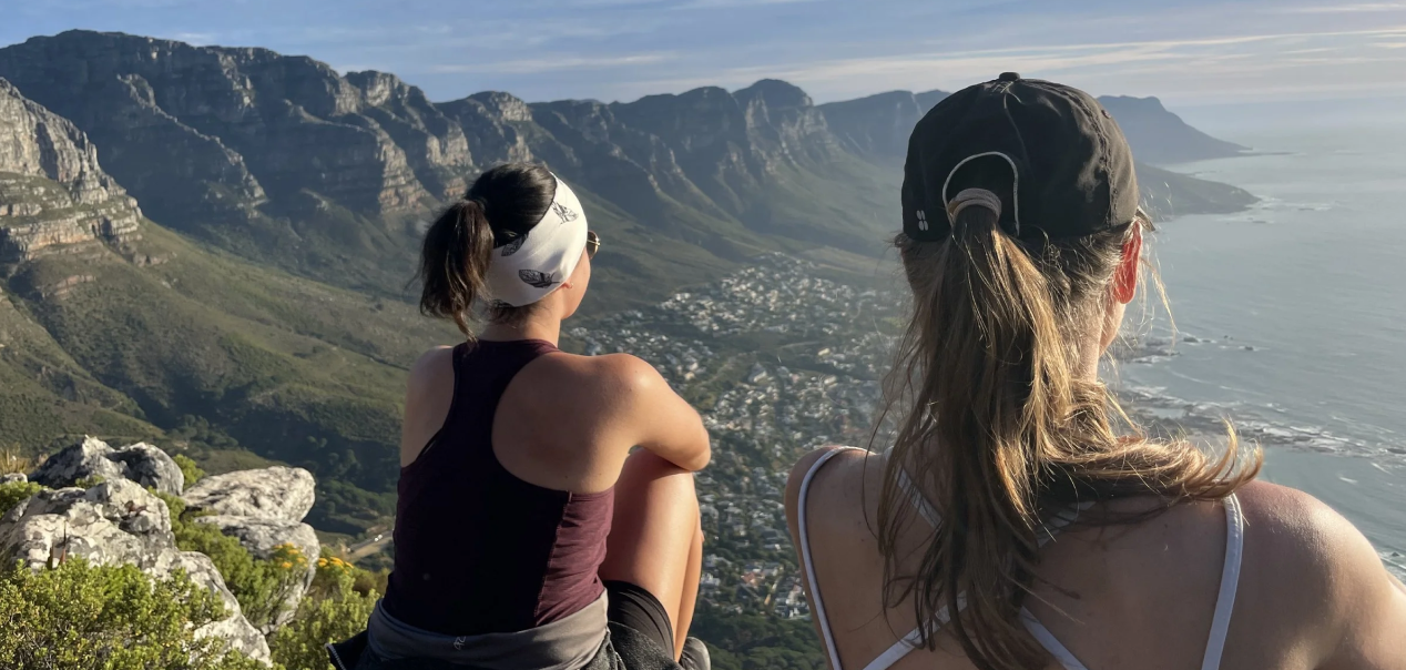 Two women sitting on a mountain overlooking a coastal town and ocean, with mountains in the background.