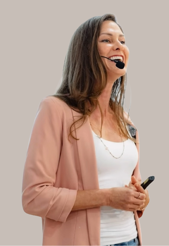 A woman with long brown hair smiling, wearing a pink blazer and white top, holding a remote control, and speaking into a headset microphone.