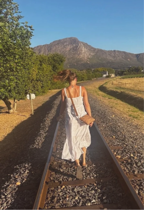 A woman walking on train tracks in a rural area with mountains in the background during daytime.
