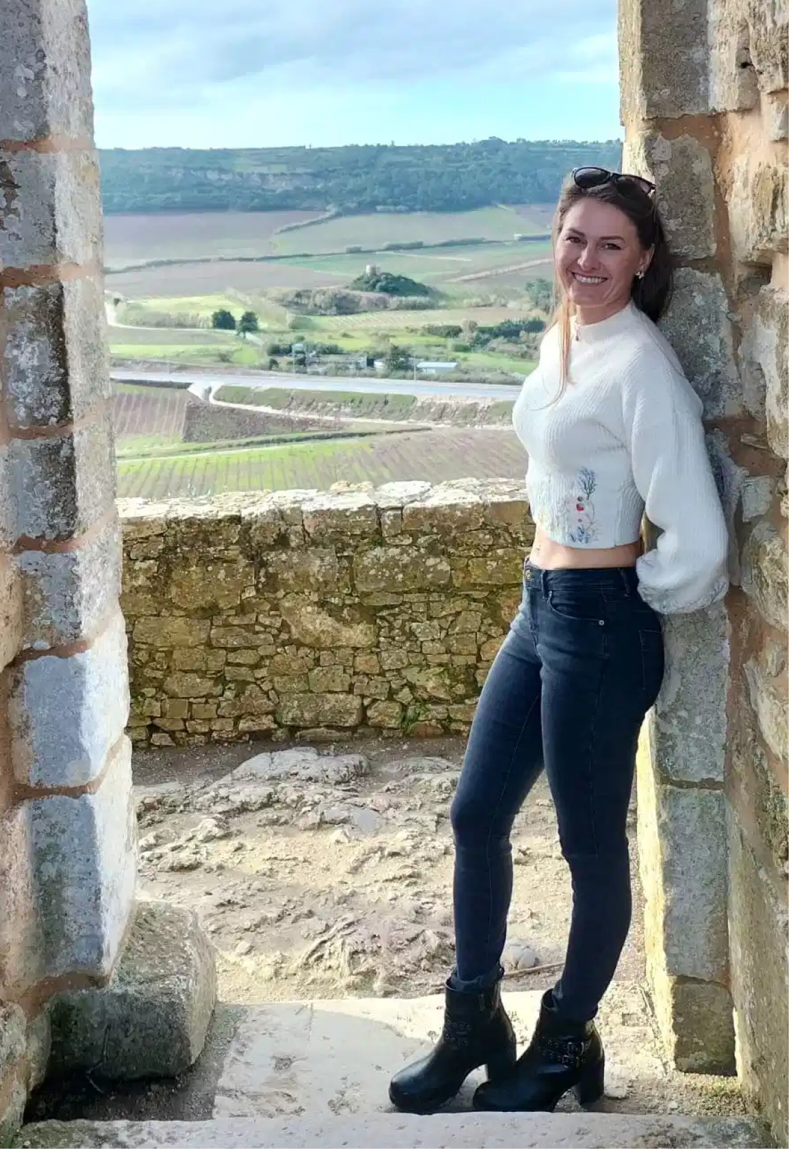 A woman leaning against a stone wall, smiling, with scenic countryside in the background.