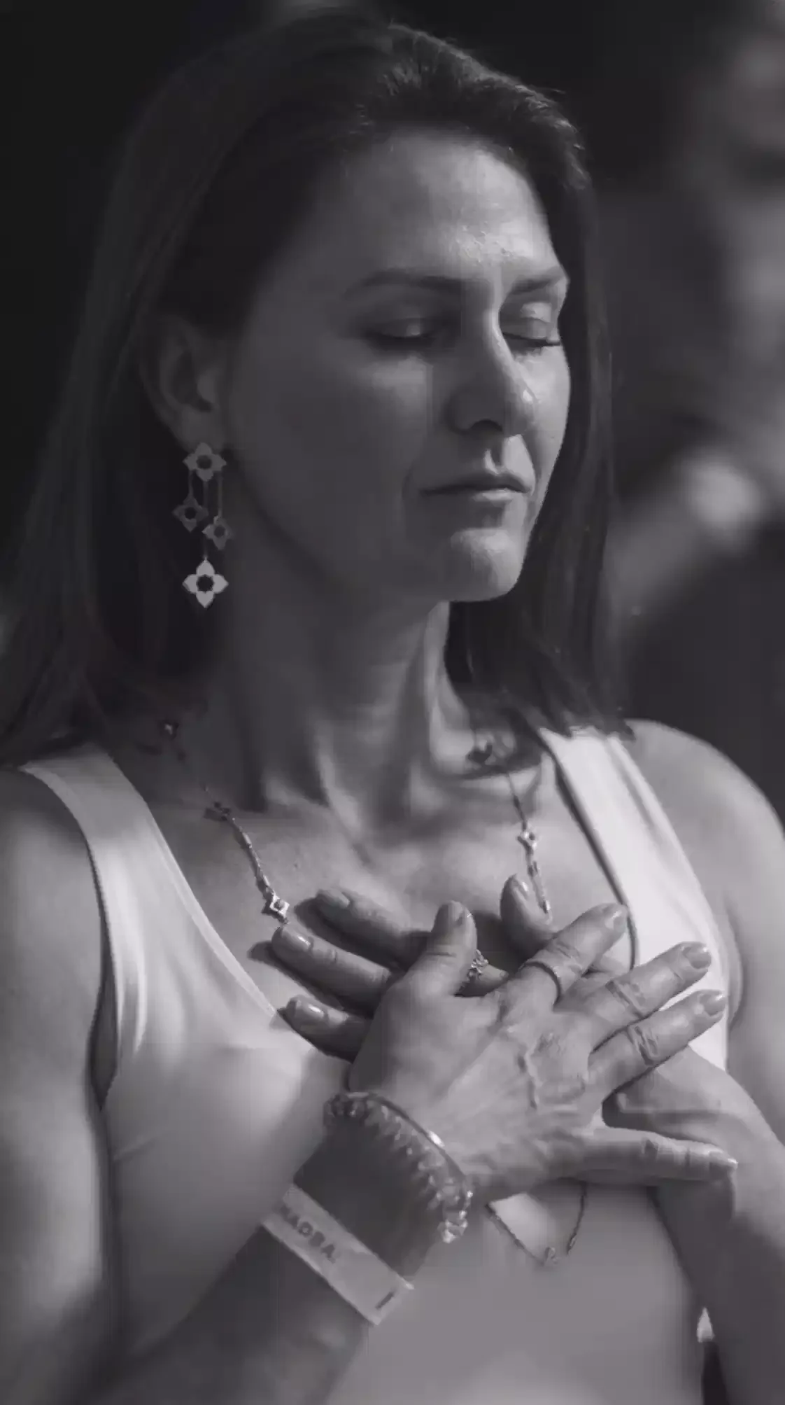 A woman with eyes closed, wearing earrings, a necklace, and a bracelet, holding her hands over her chest with a serene expression.