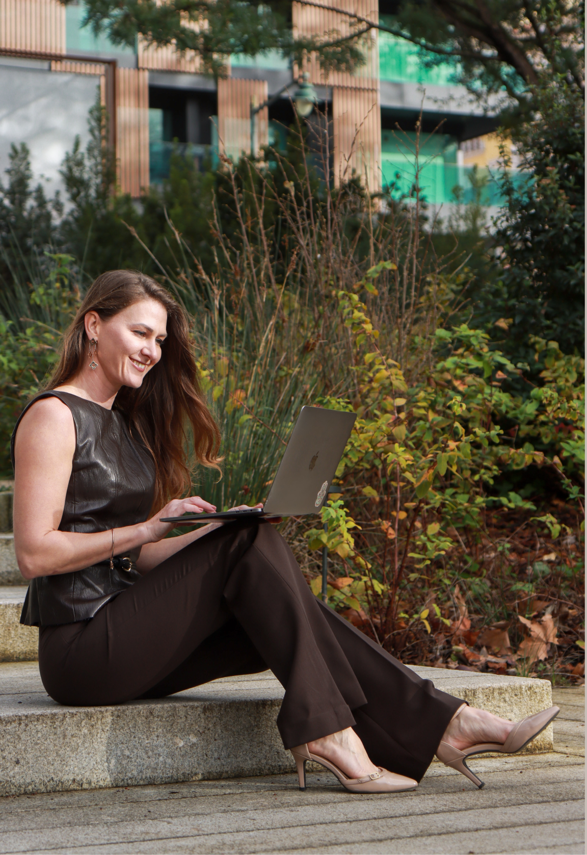 A woman sitting on outdoor stairs using a laptop, wearing a black leather top, brown pants, and beige high heels, with greenery and modern buildings in the background.