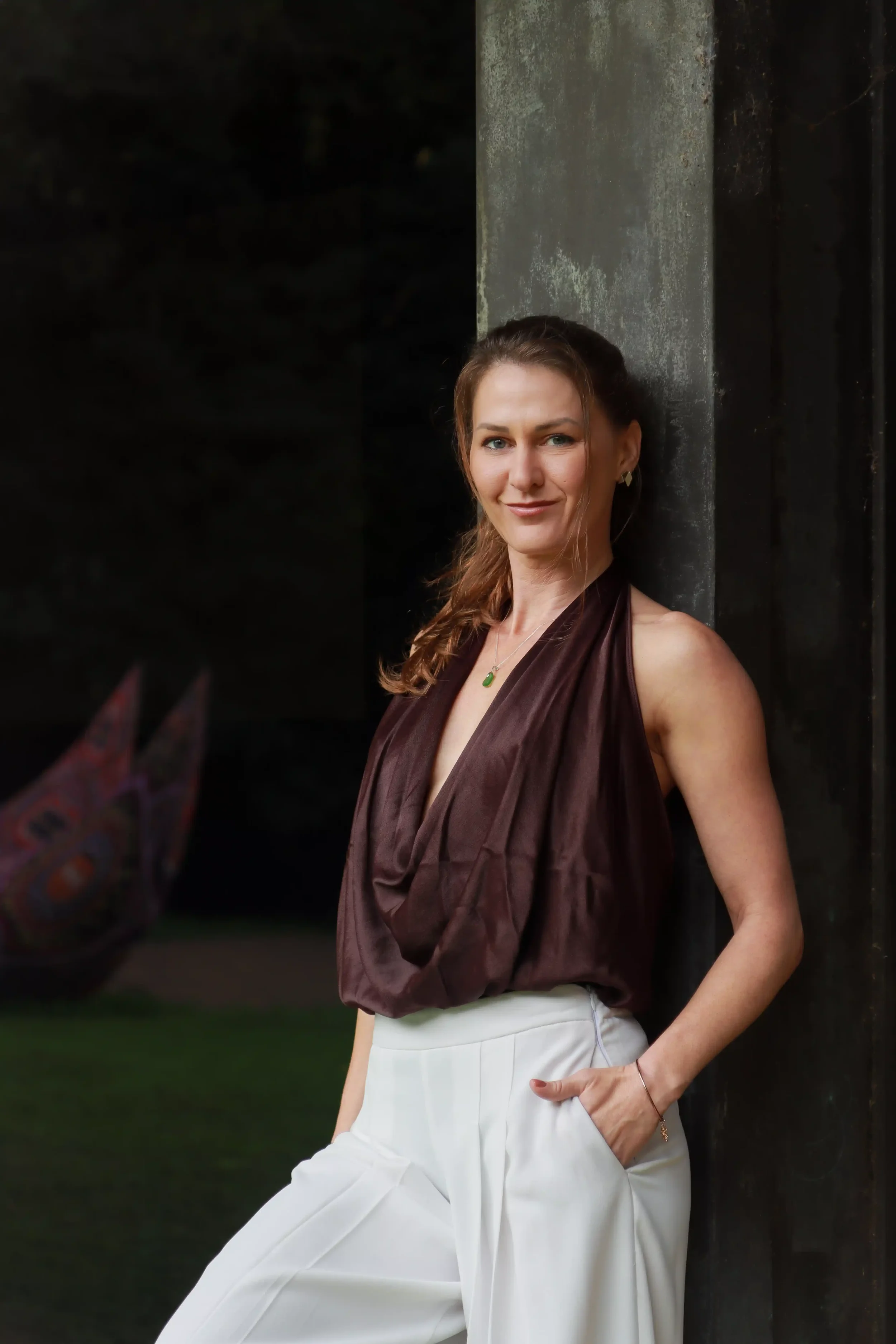 Woman with light skin, brown hair, wearing a silky brown top and white pants, standing outdoors against a dark background and concrete pillar, smiling at the camera.