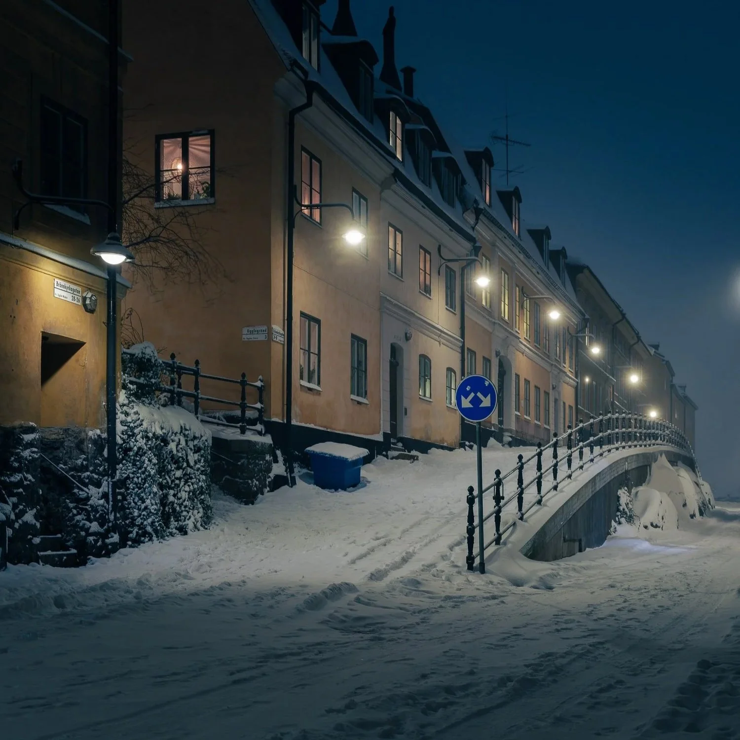 Snow-covered street with residential buildings at night. Street lamps illuminate the area, and a bridge crosses over a frozen body of water. Windows in the buildings emit a warm glow.