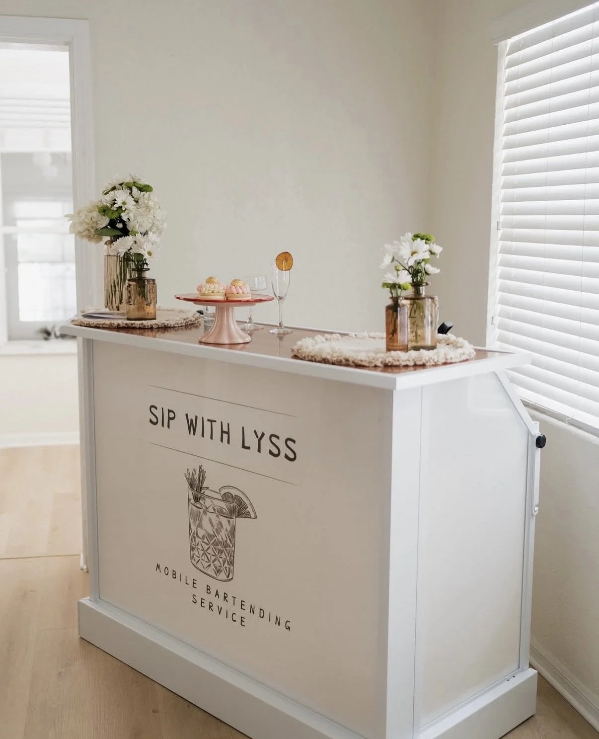 A white mobile bartending service cart with flowers and desserts on top, decorated with a sign that reads 'Sip with Lyss' and an illustration of a cocktail glass.