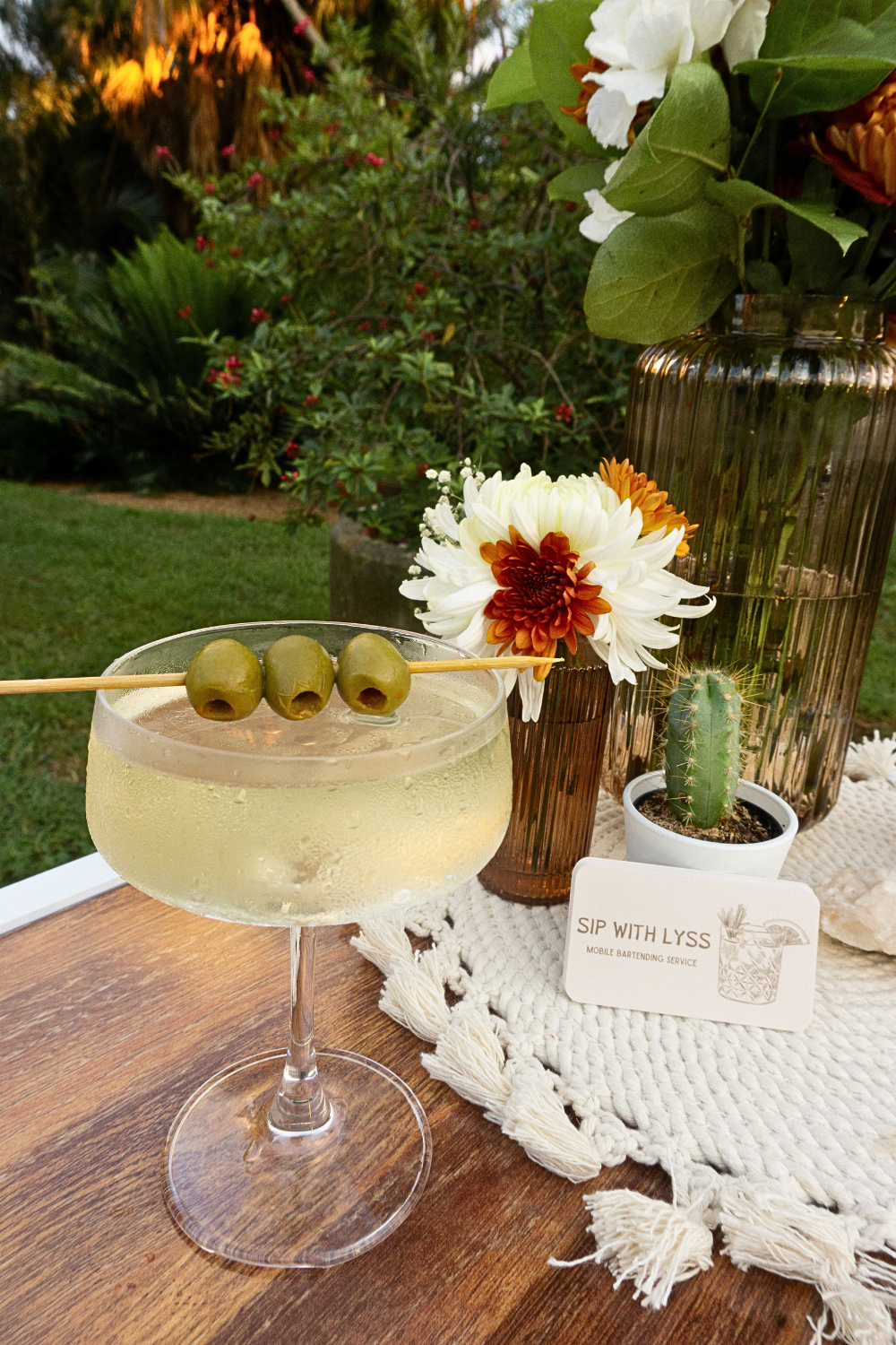 A cocktail garnished with three green olives served on a wooden table outdoors. Decorative flowers and a small cactus in a white pot are arranged beside the drink, along with a business card that reads 'Sip with Lyss, Mobile Bartending Service'.