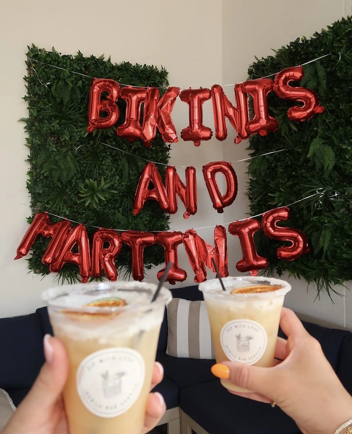 Two people holding drinks in front of a Christmas-themed celebration with a red balloon banner reading 'Bikini's and Martinis' and a green leafy backdrop.