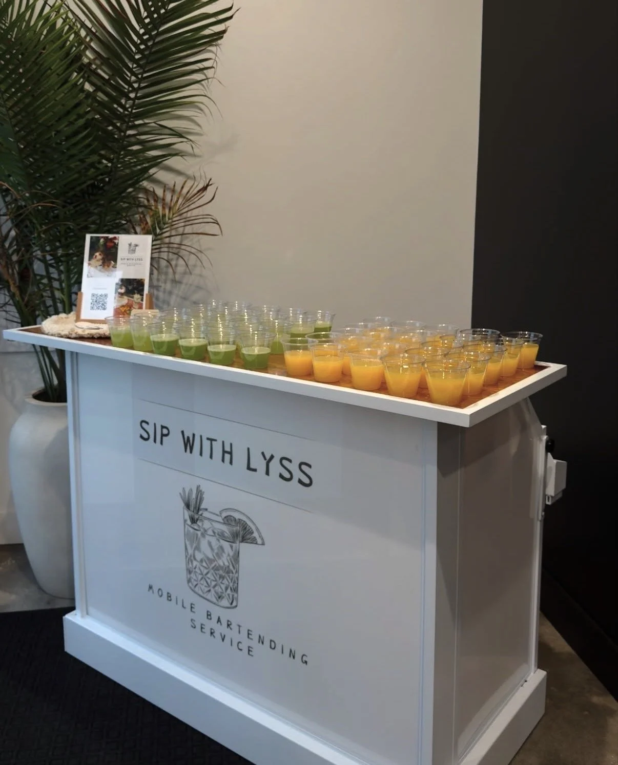 A mobile bartending service setup featuring a white cart with the slogan 'Sip with Lyss' and a graphic of a drink glass. On top of the cart, there are rows of green and orange drinks in clear cups. To the left, a large potted plant and a small stand 
