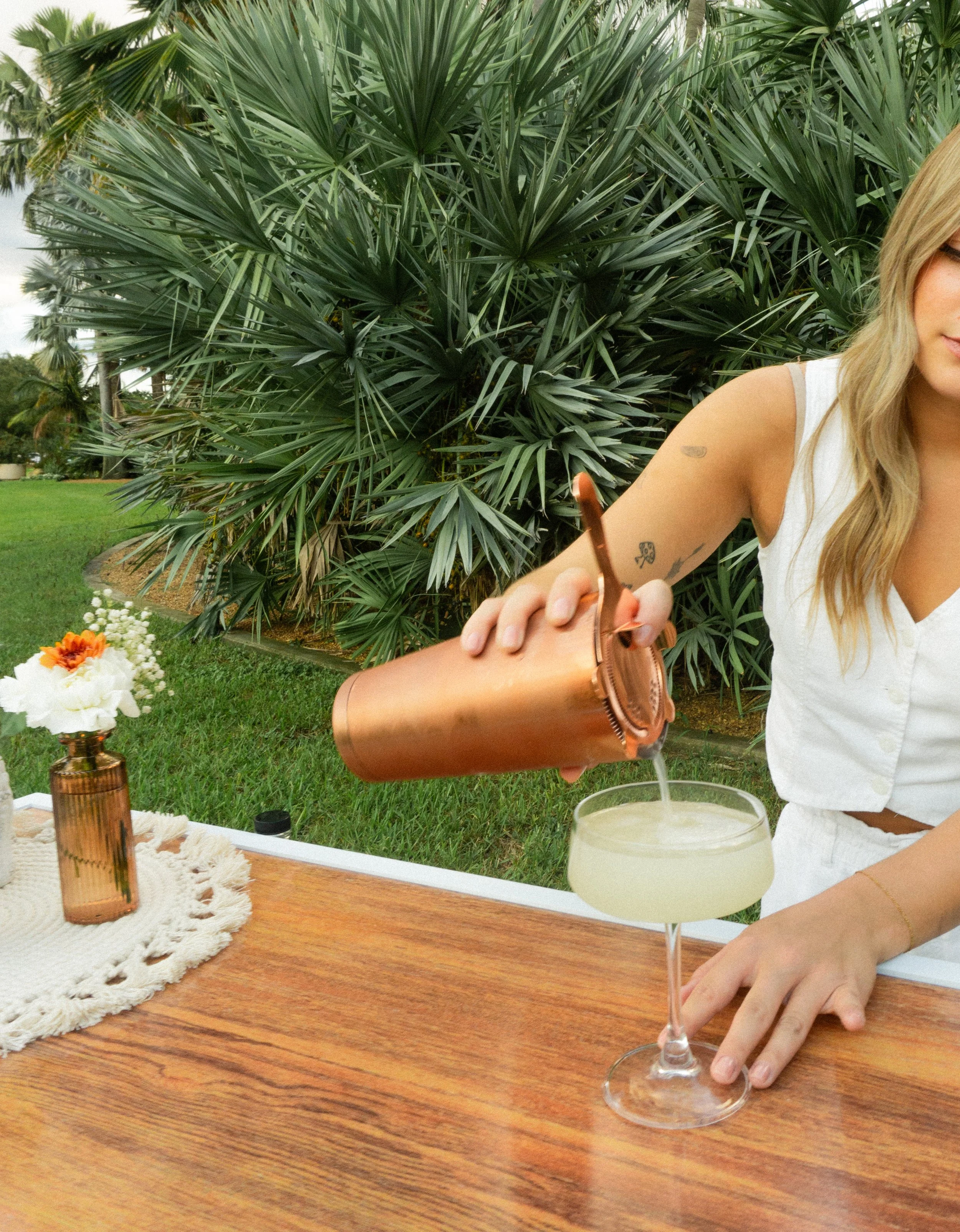 A woman pouring a cocktail from a copper shaker into a coupe glass outdoors at a garden party. There is a small flower arrangement on a white tablecloth nearby.