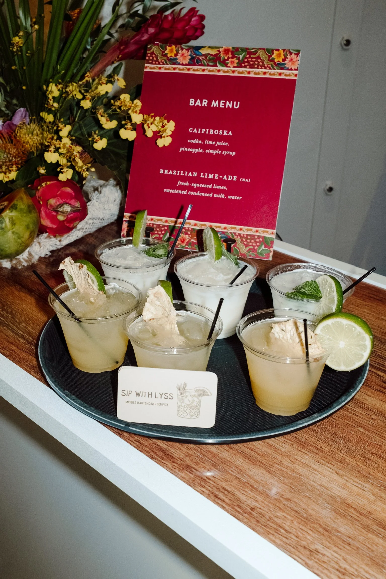 Six pre-mixed cocktails with lime wedges and ice on a black tray, placed on a wooden surface. A floral sign and a colorful banner are visible in the background, along with some flowers.