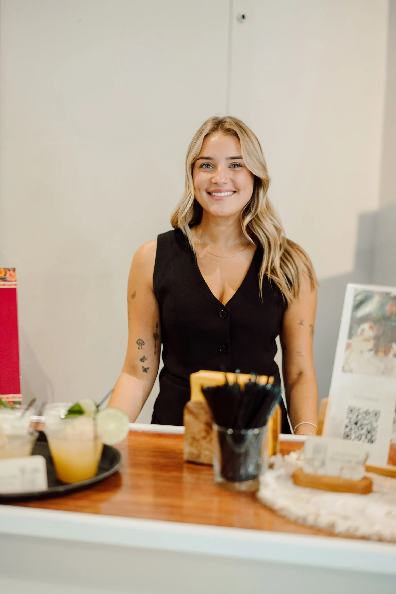 A smiling blonde woman standing behind a counter at a cafe or restaurant, wearing a sleeveless black top with tattoos on her arms, with drinks and supplies on the counter in front of her.