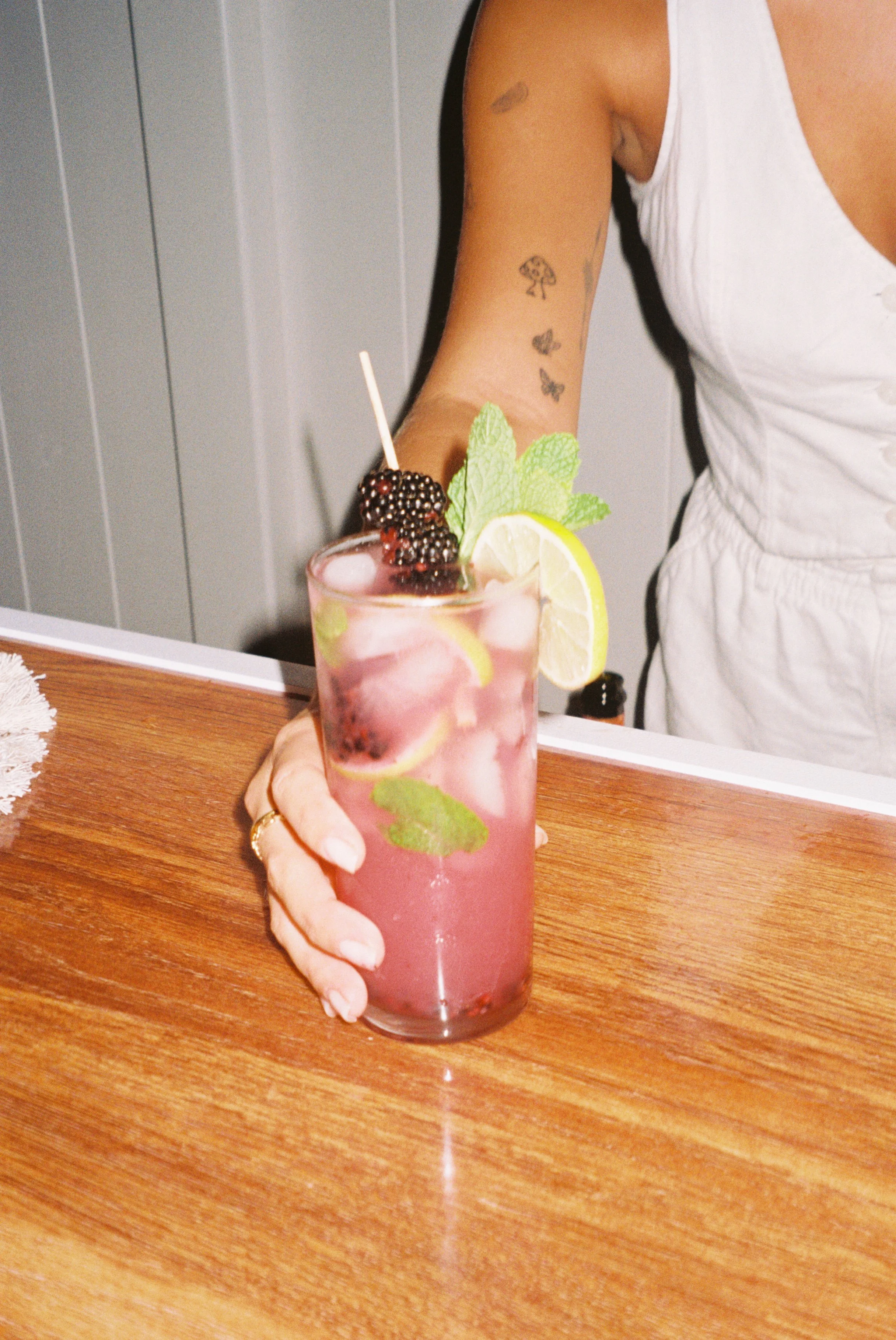 A woman with tattoos on her arm holding a pink cocktail garnished with lemon slices, blackberries, mint leaves, and a skewer with berries.