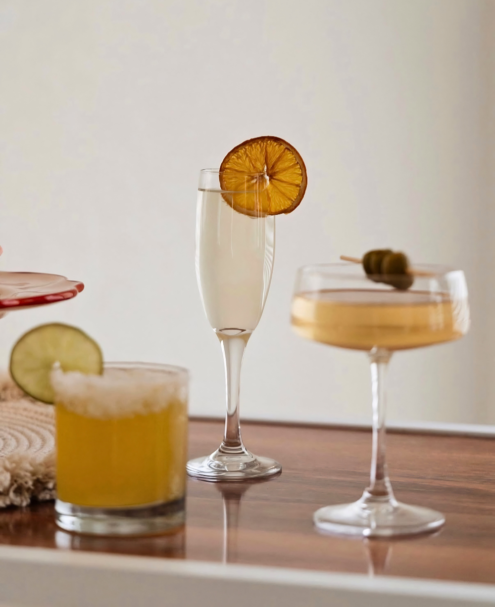 Four different cocktails on a wooden surface, each garnished with fruit slices or olives, with a plain white wall in the background.