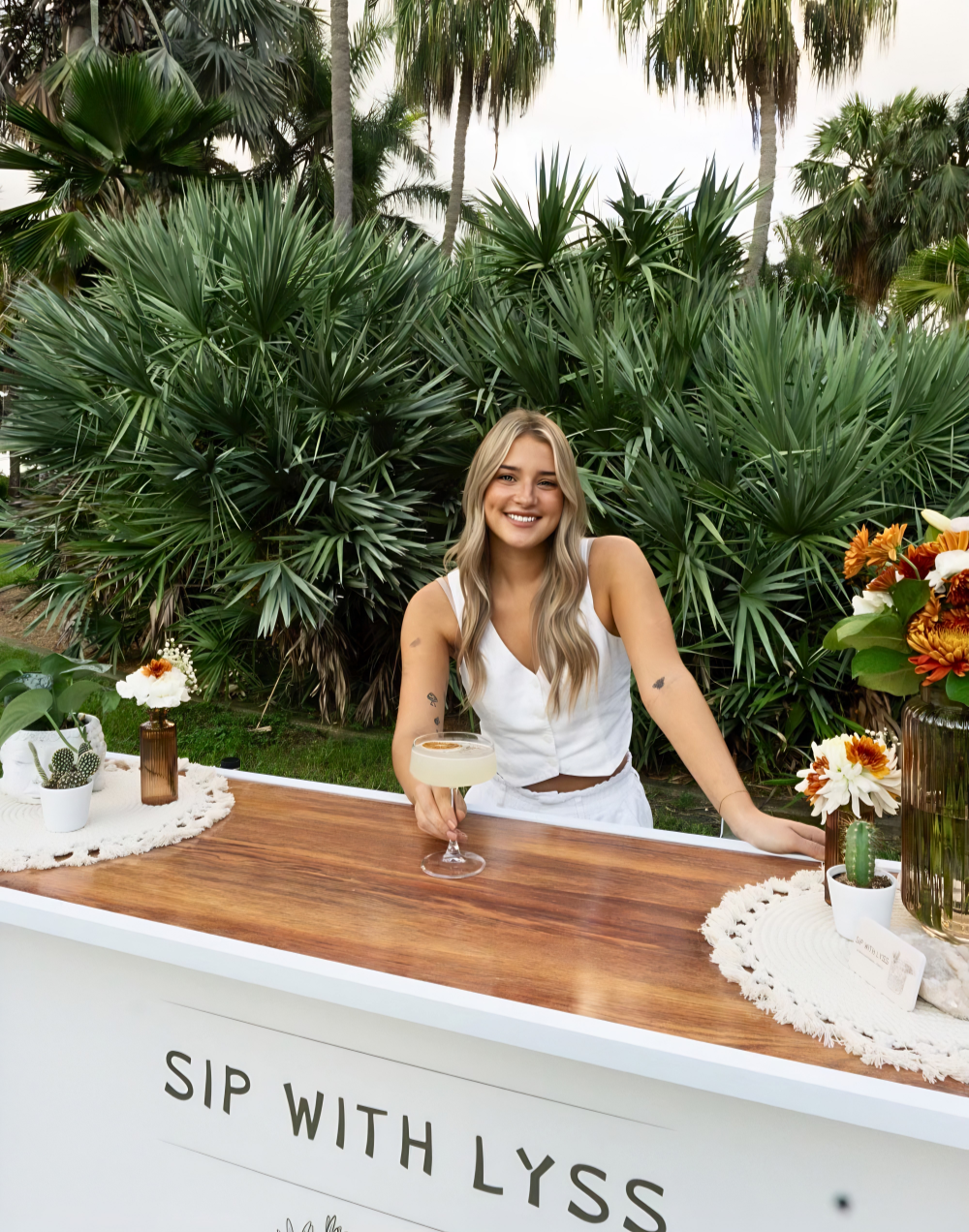 A woman standing behind a bar with tropical plants in the background, holding a cocktail, and smiling.