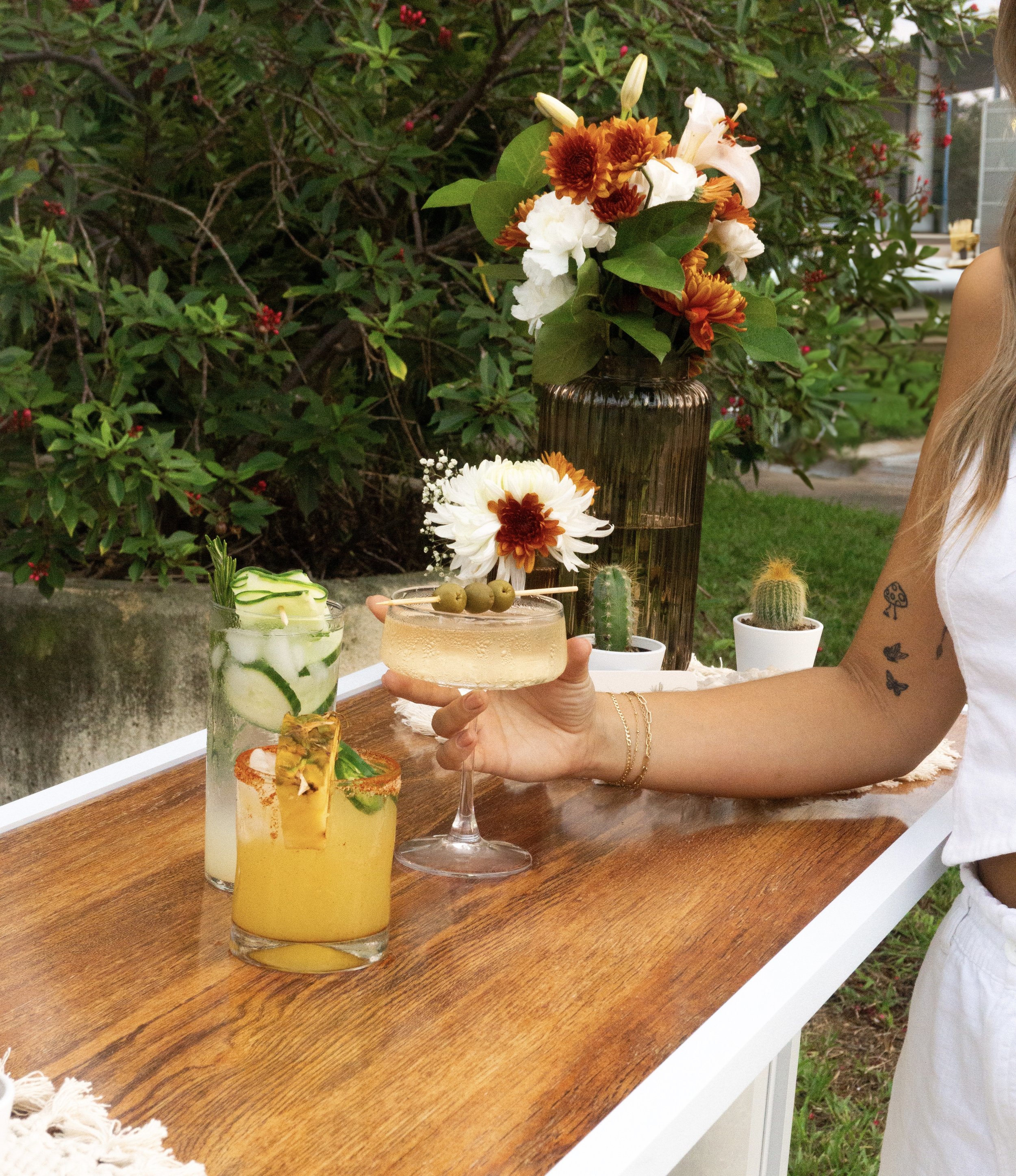 A person holding a light-colored cocktail with an olive garnish over a wooden table with two other cocktails, one with cucumber slices and the other with pineapple and lime, on a patio with potted cacti and a large flower arrangement in a vase.