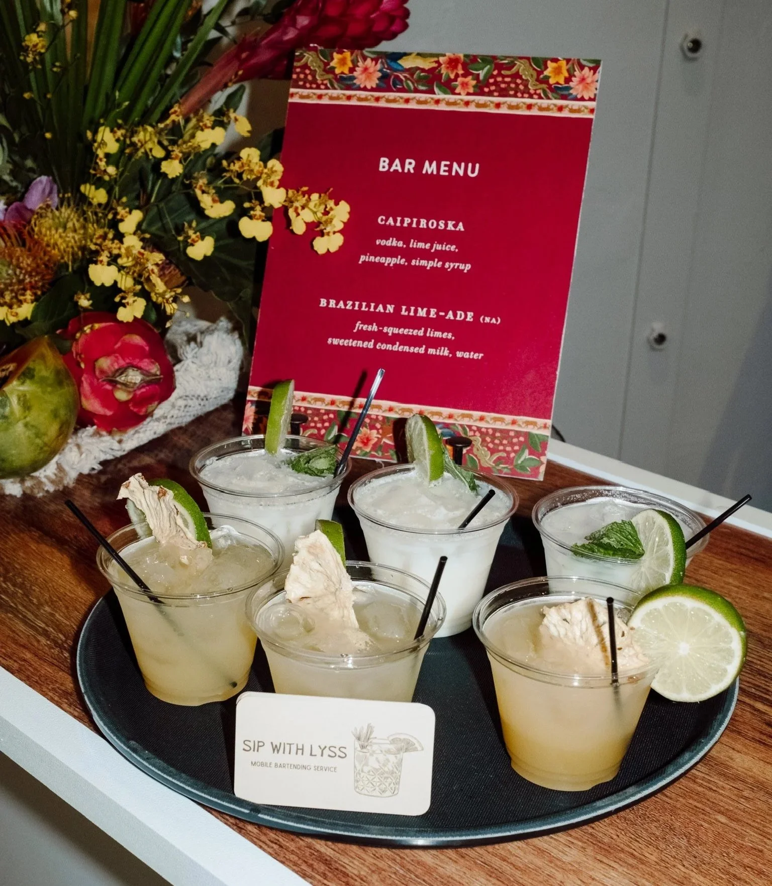 Collection of five cocktails with lime wedges and mint leaves on a tray, behind a red printed menu with floral decorative borders listing drinks called Caipiroska and Brazilian Lime-Ade, on a wooden surface with flowers in the background.