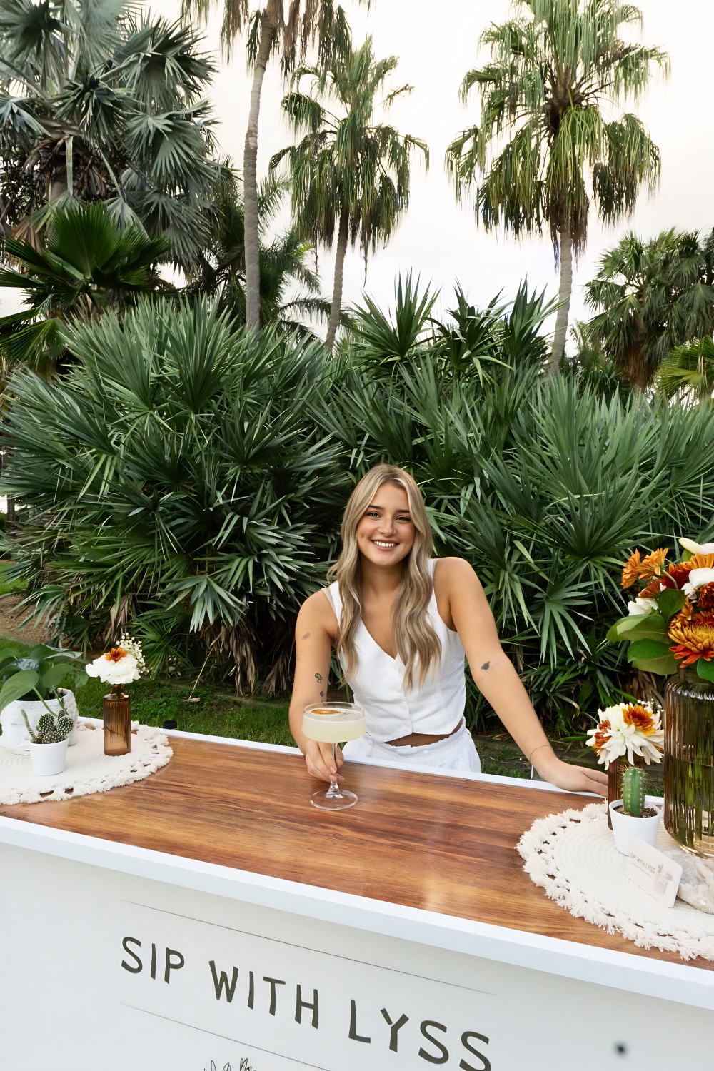 A smiling young woman with long blonde hair and tattoos on her arms stands behind a white bar with a wooden top outdoors, surrounded by large green tropical plants and tall palm trees, holding a cocktail glass with a light-colored drink.