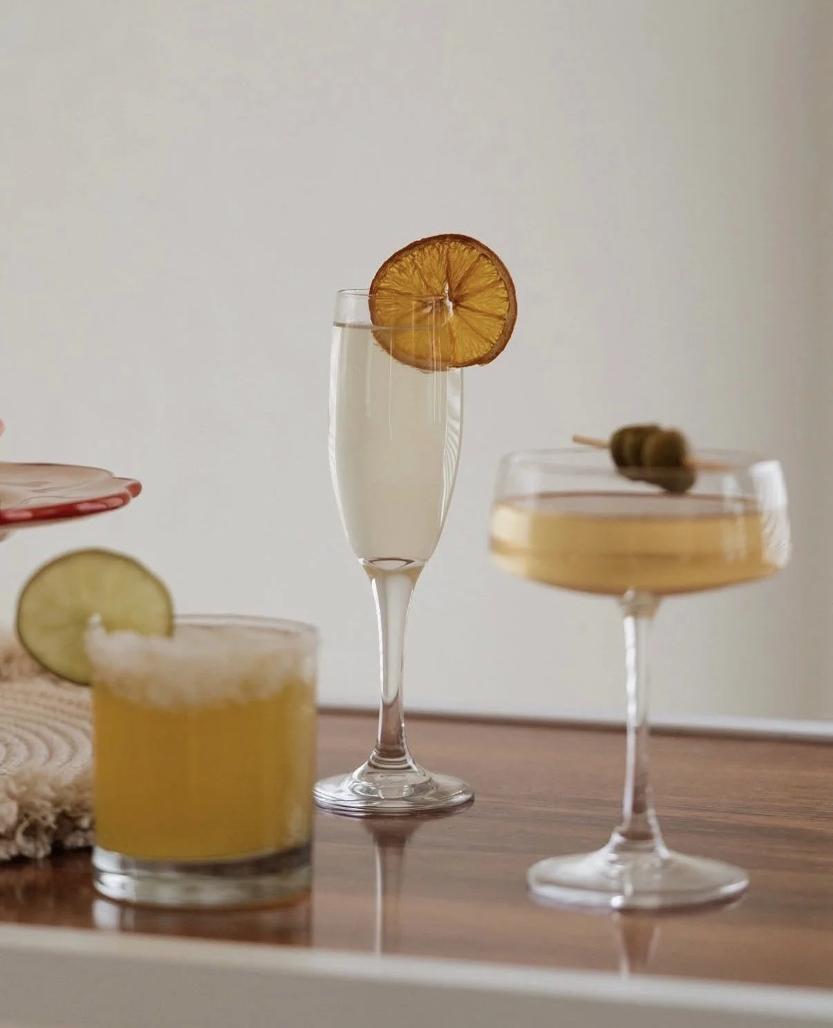 Various cocktails and drinks on a wooden surface, including a champagne flute with a lemon slice, a short glass with a yellow beverage and lemon garnish, and a coupe glass with a light-colored drink and an olive skewer.