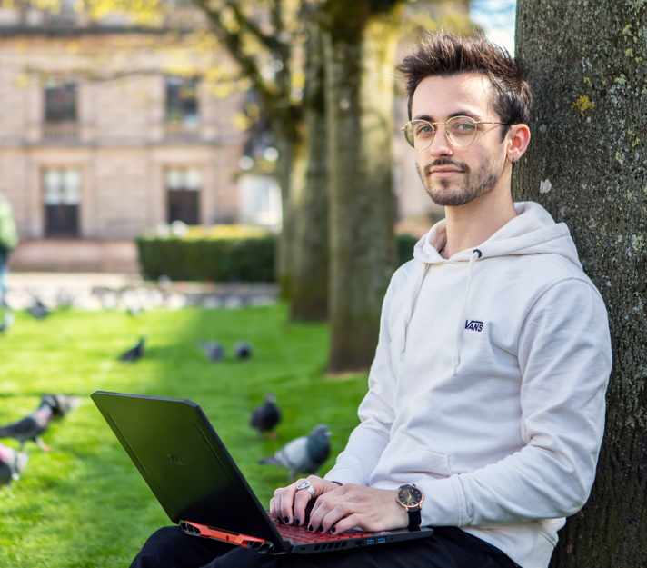 Young man with glasses and a white hoodie sitting against a tree in a park, using a laptop with pigeons in the background.