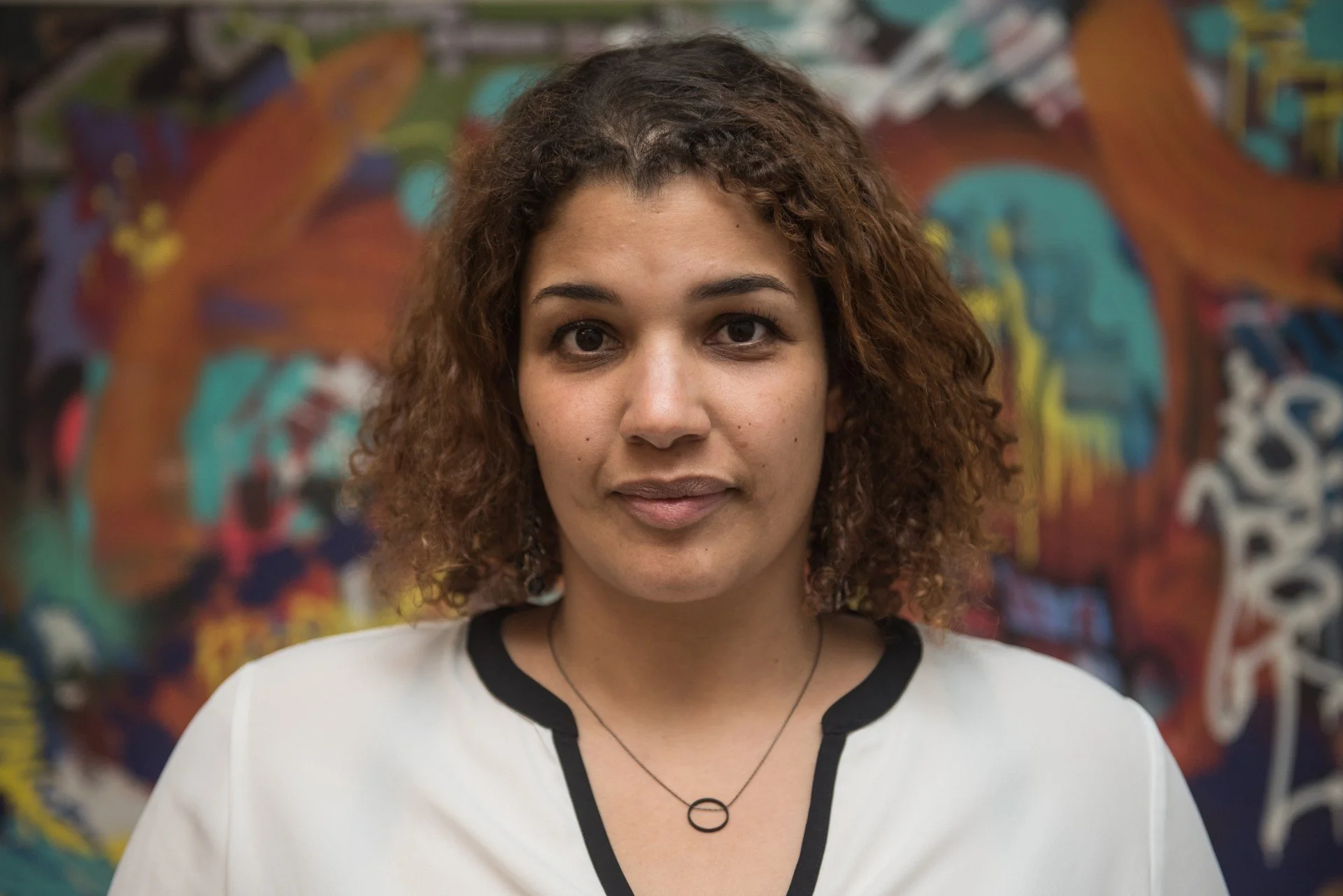 Jeune femme avec bouclés marron, portant un t-shirt blanc avec col noir, devant un mur avec graffitis colorés.
