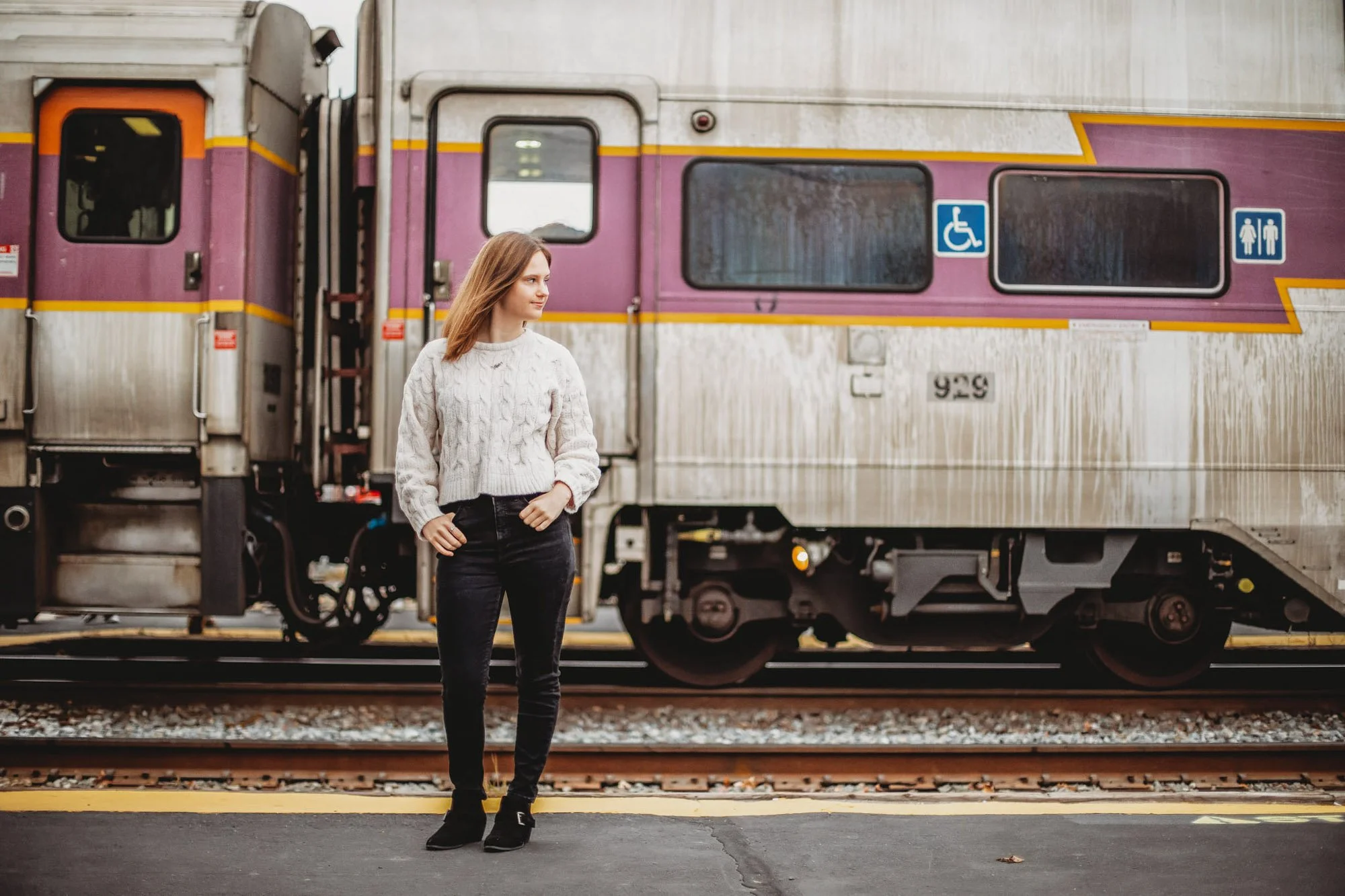 A young woman in a white sweater and black pants standing on train tracks at a train station, with a purple and gray train in the background.
