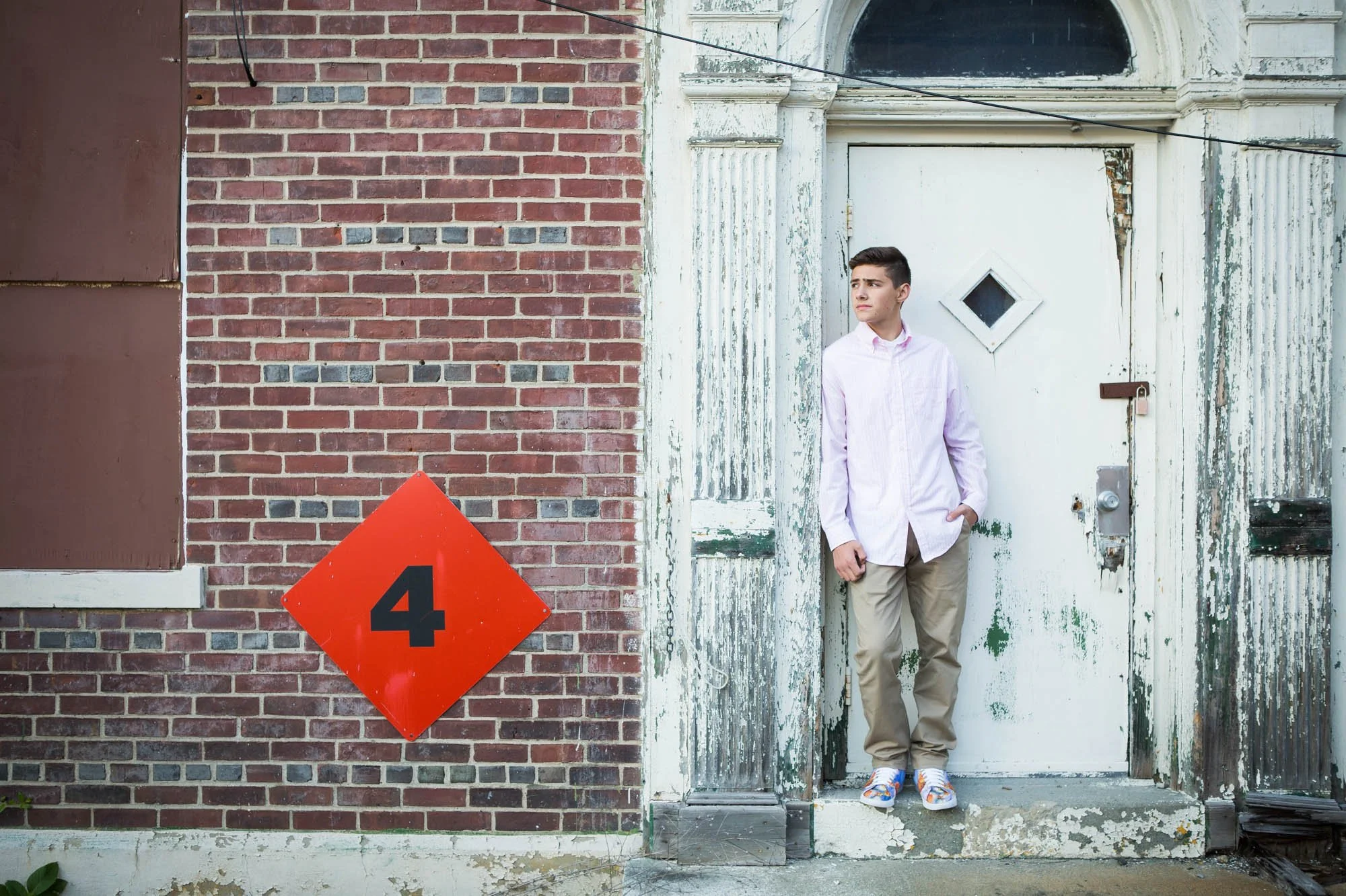 Young man with short dark hair wearing a light pink shirt, beige pants, and white sneakers standing at an old white door with peeling paint, next to a brick wall with a red diamond-shaped sign displaying the number 4.