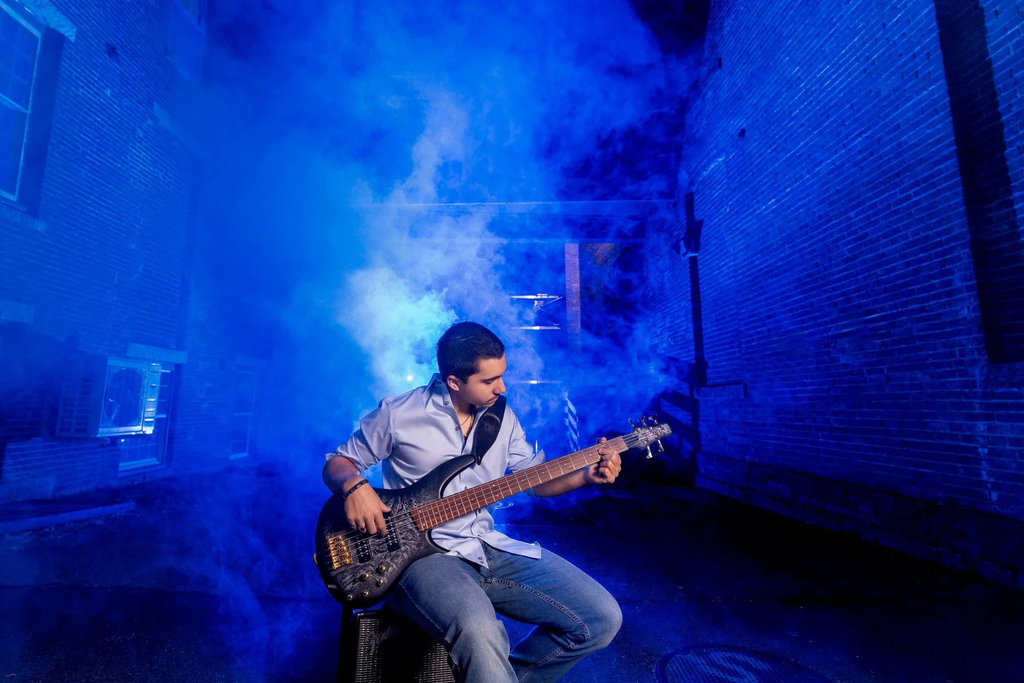 A young man is sitting on a small amplifier, playing an electric bass guitar in a dimly lit alleyway with blue lighting and smoke, flanked by brick buildings.