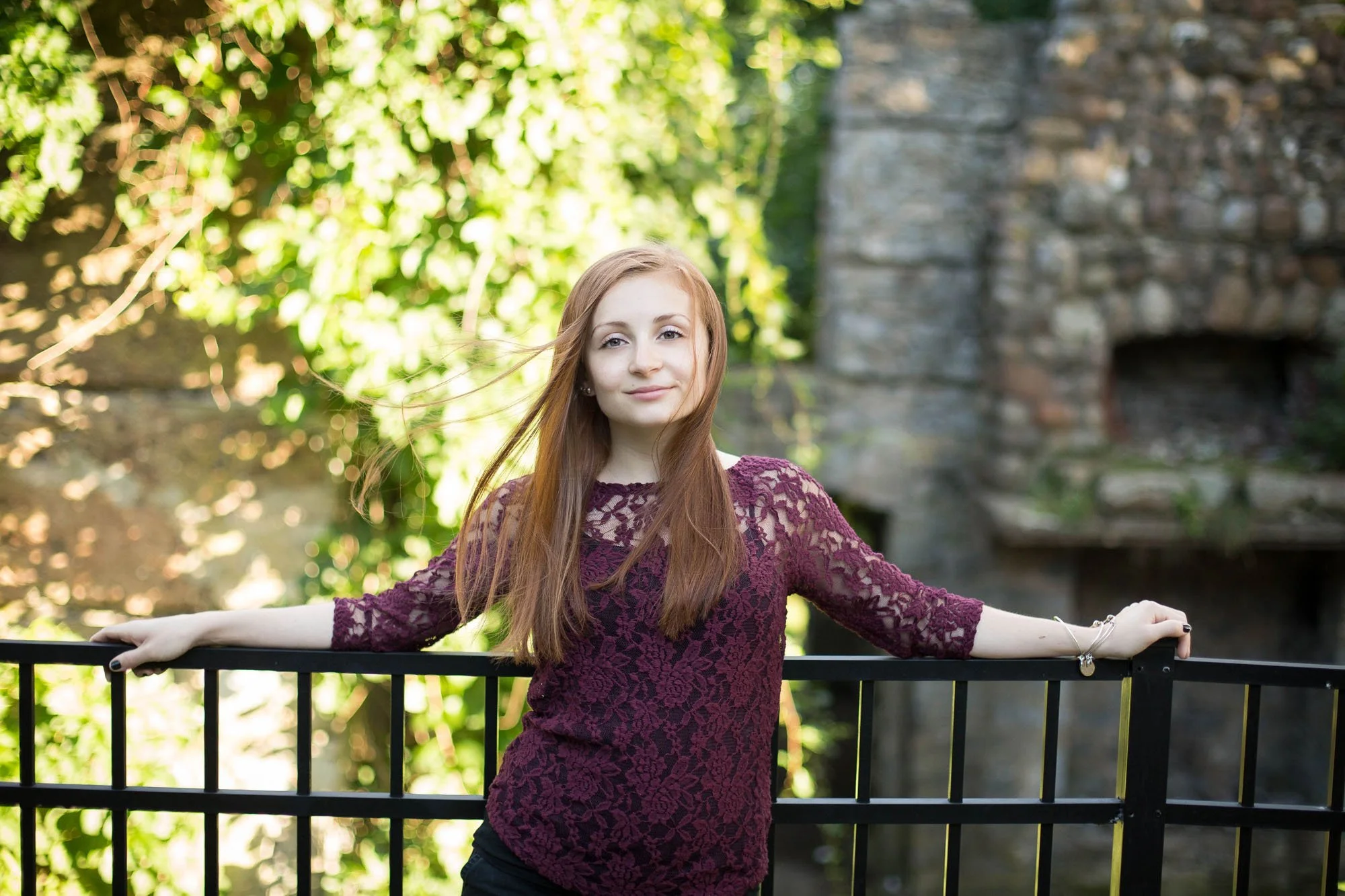 A young woman with long red hair wearing a burgundy lace top, standing outdoors near a black metal fence with greenery and a stone structure in the background.