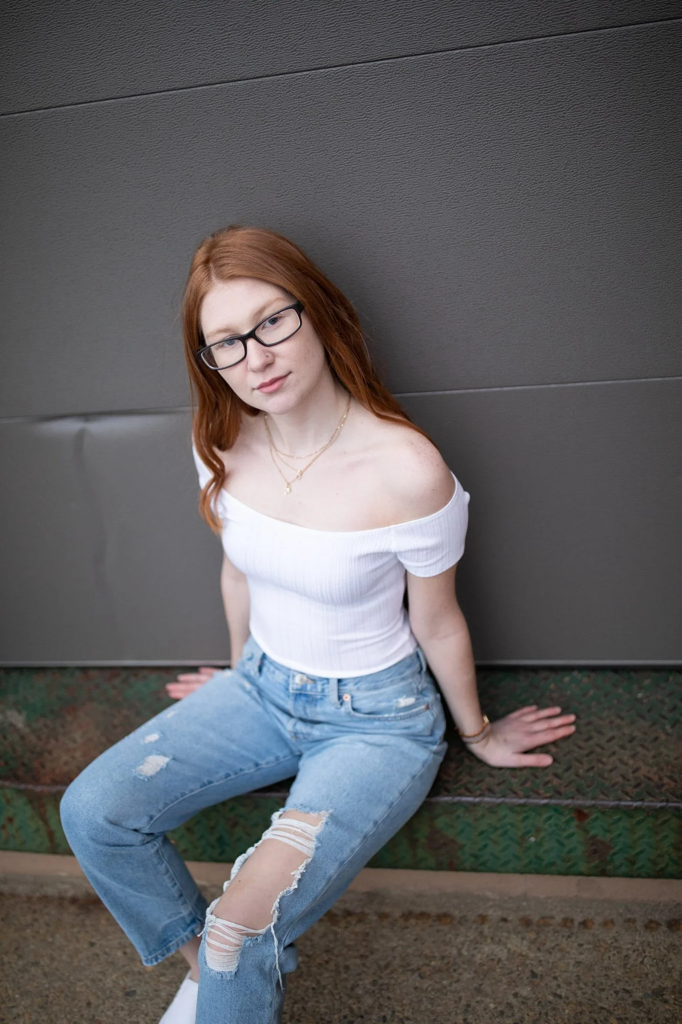 A young woman with red hair and glasses sitting on a green metal surface against a dark gray wall, wearing a white off-shoulder top and ripped blue jeans.