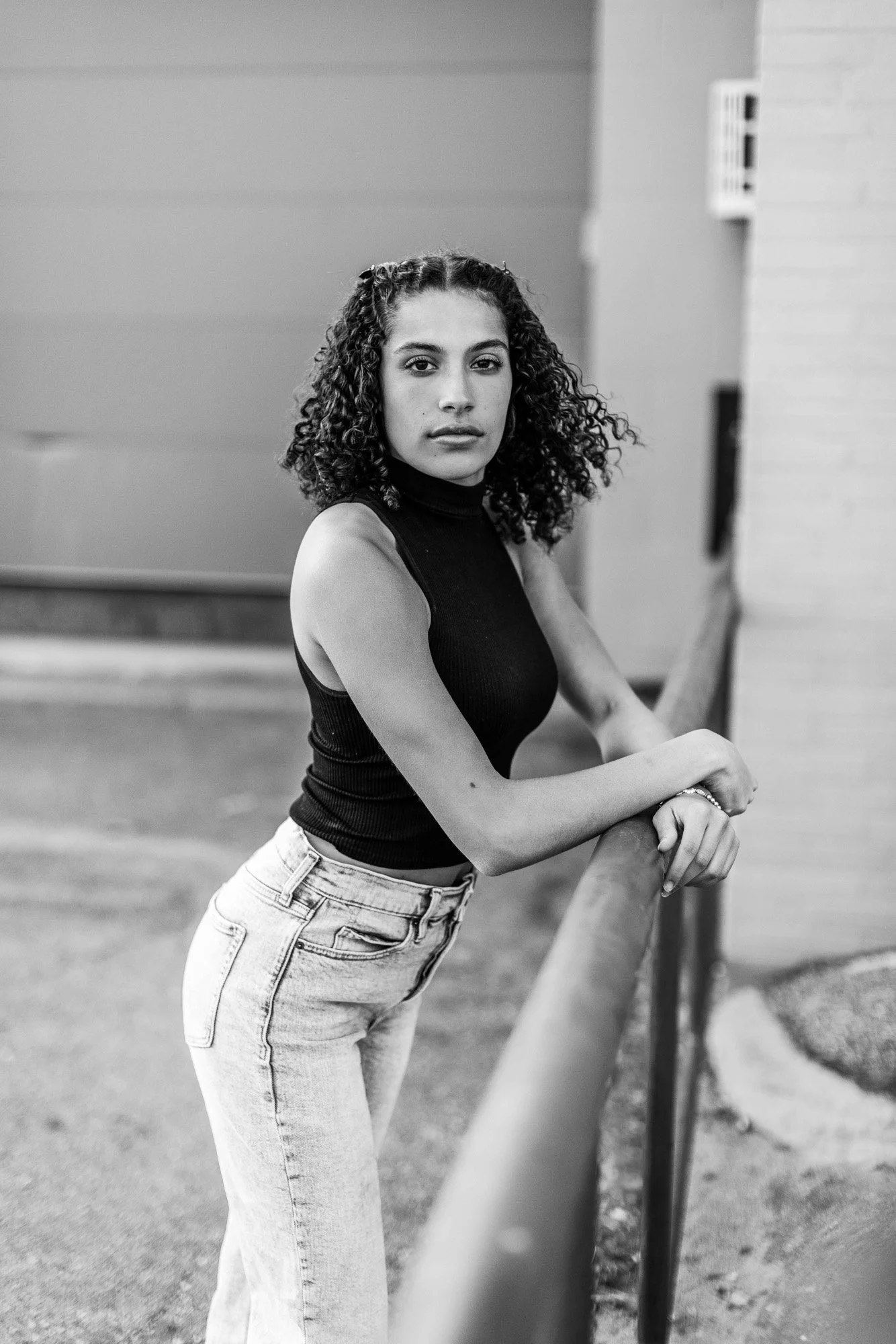A black and white photo of a young woman with curly hair, wearing a sleeveless black top and light-colored jeans, leaning on a railing and looking at the camera.