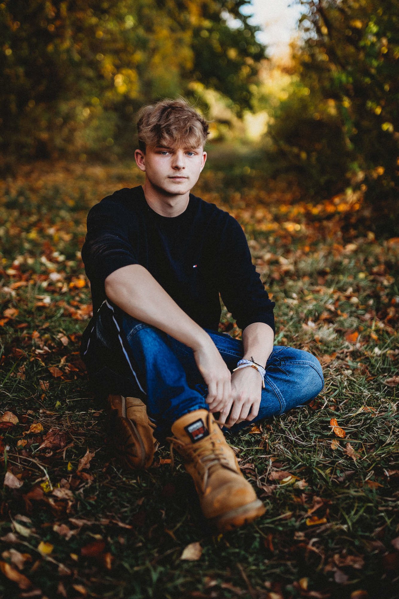 A young man with light brown hair and fair skin sitting on autumn leaves in a forested area, wearing a black long-sleeve shirt, blue jeans, and tan work boots.