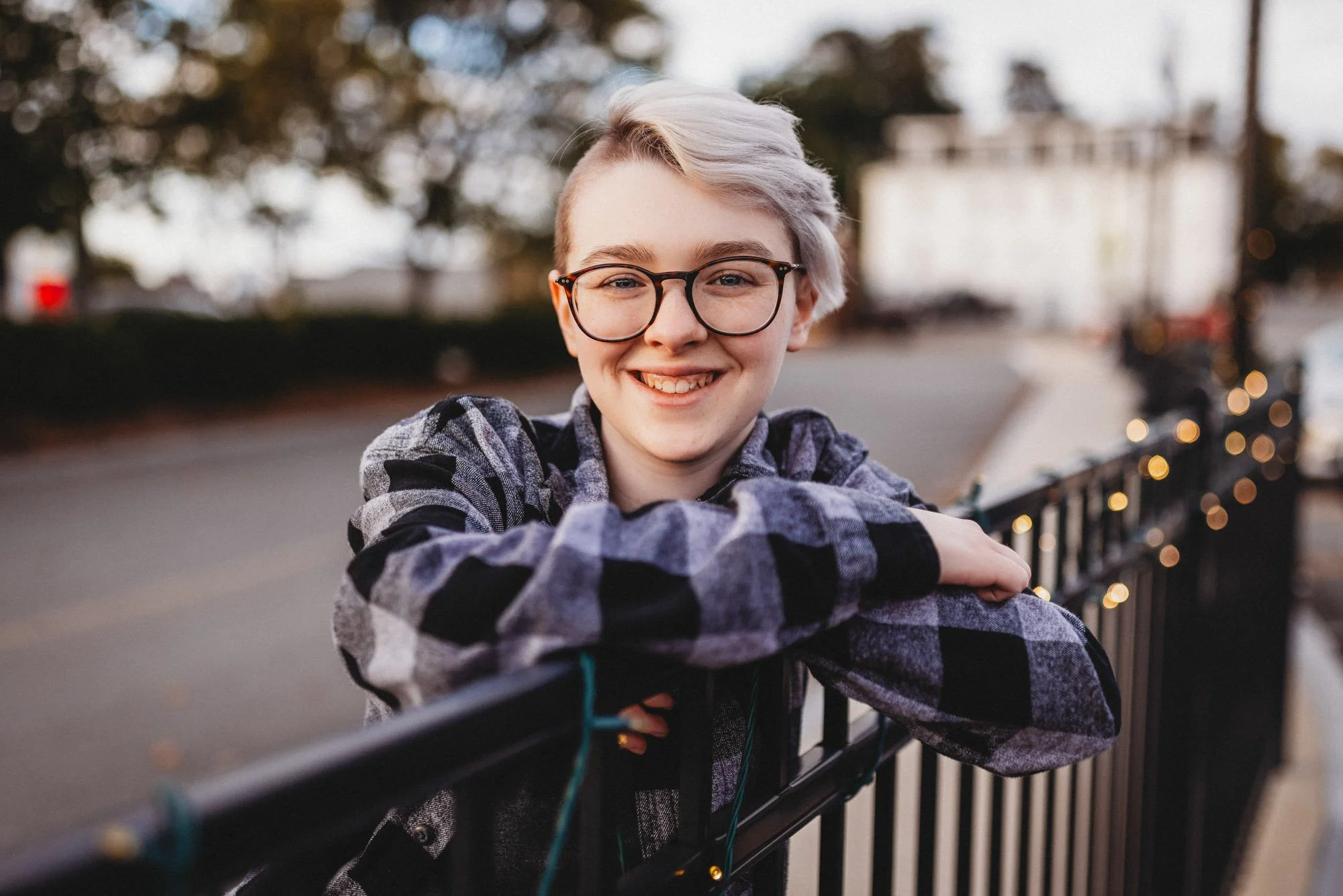 Smiling young person with glasses and short, light-colored hair leaning on a black metal fence decorated with string lights outdoors.