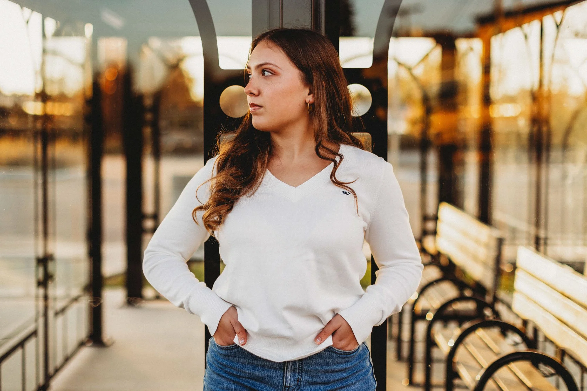 Young woman with long curly hair, wearing a white sweater and blue jeans, standing at a bus stop with her hands in her pockets, during sunset.