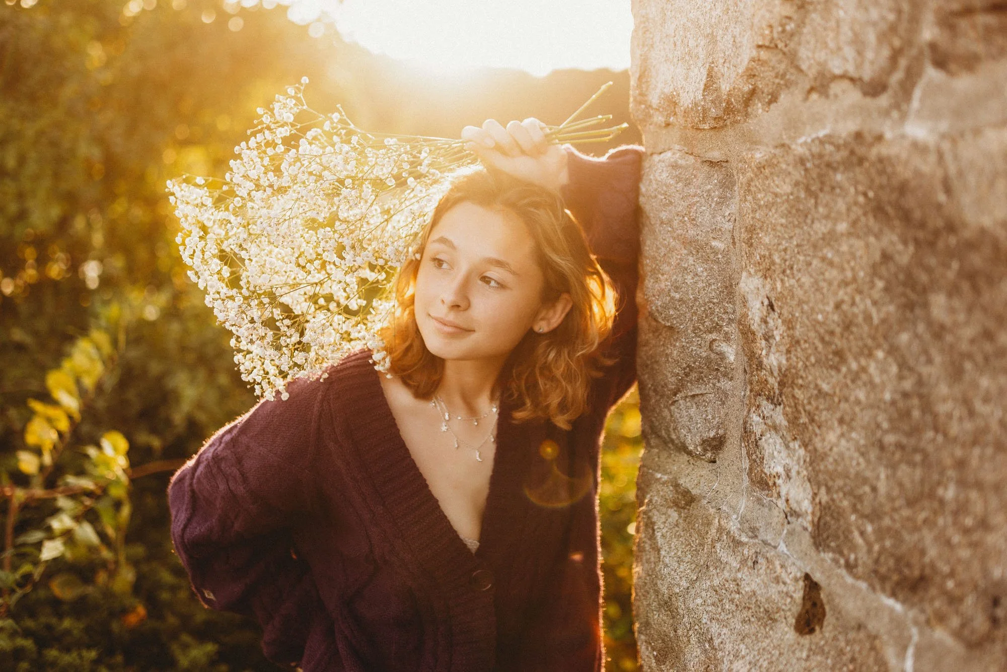 A young woman with curly red hair leaning against a stone wall, holding a bouquet of white flowers, and gazing away with a peaceful expression as the sunset backlights the scene.
