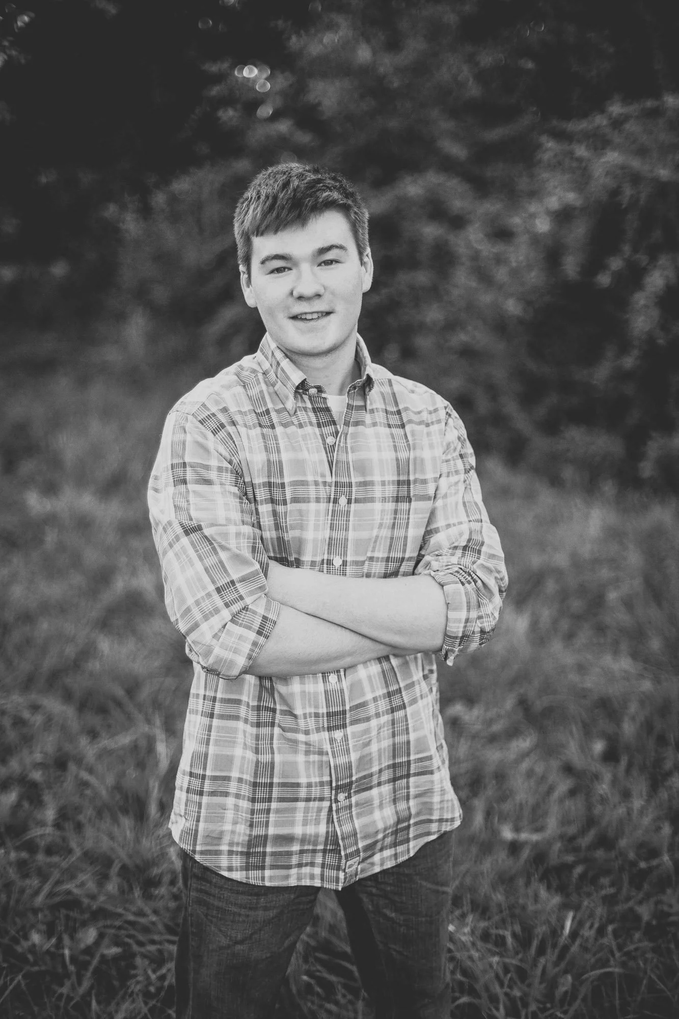 A young man with short hair and a checkered shirt standing outdoors with arms crossed, smiling at the camera, with trees and grass in the background, in black and white.
