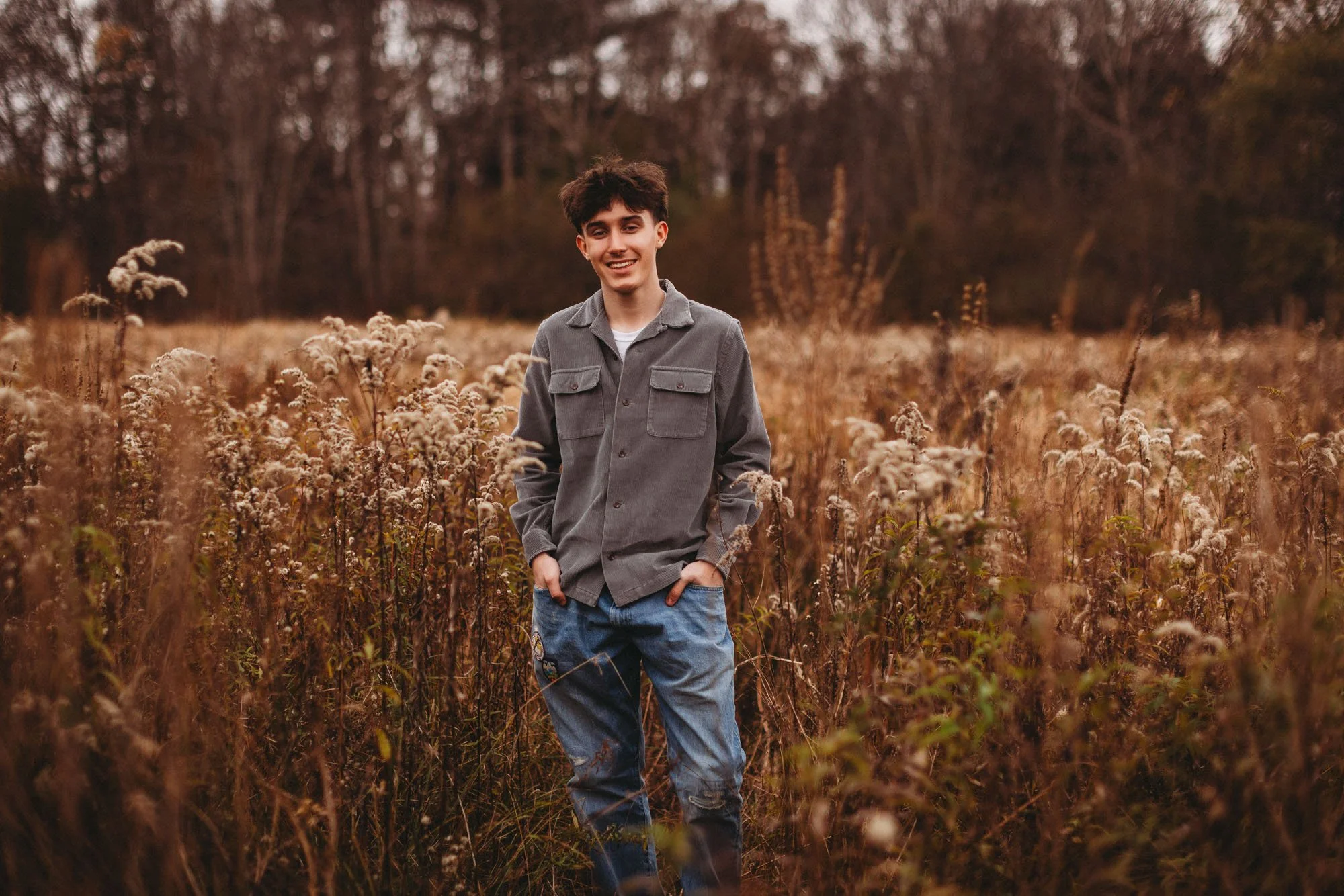 A young man with dark, tousled hair stands in a field of tall, dried grass and wildflowers, smiling at the camera. He is wearing a gray button-up shirt and ripped blue jeans, with hands in his pockets. The background features a line of trees with aut