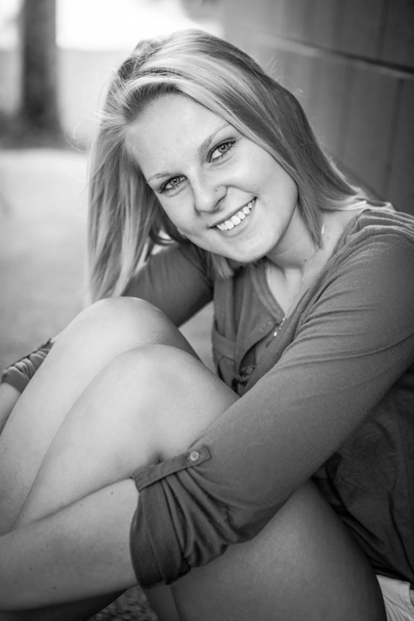 Black and white photo of a smiling young woman with shoulder-length hair, sitting outdoors with her knees up, leaning against a wall.