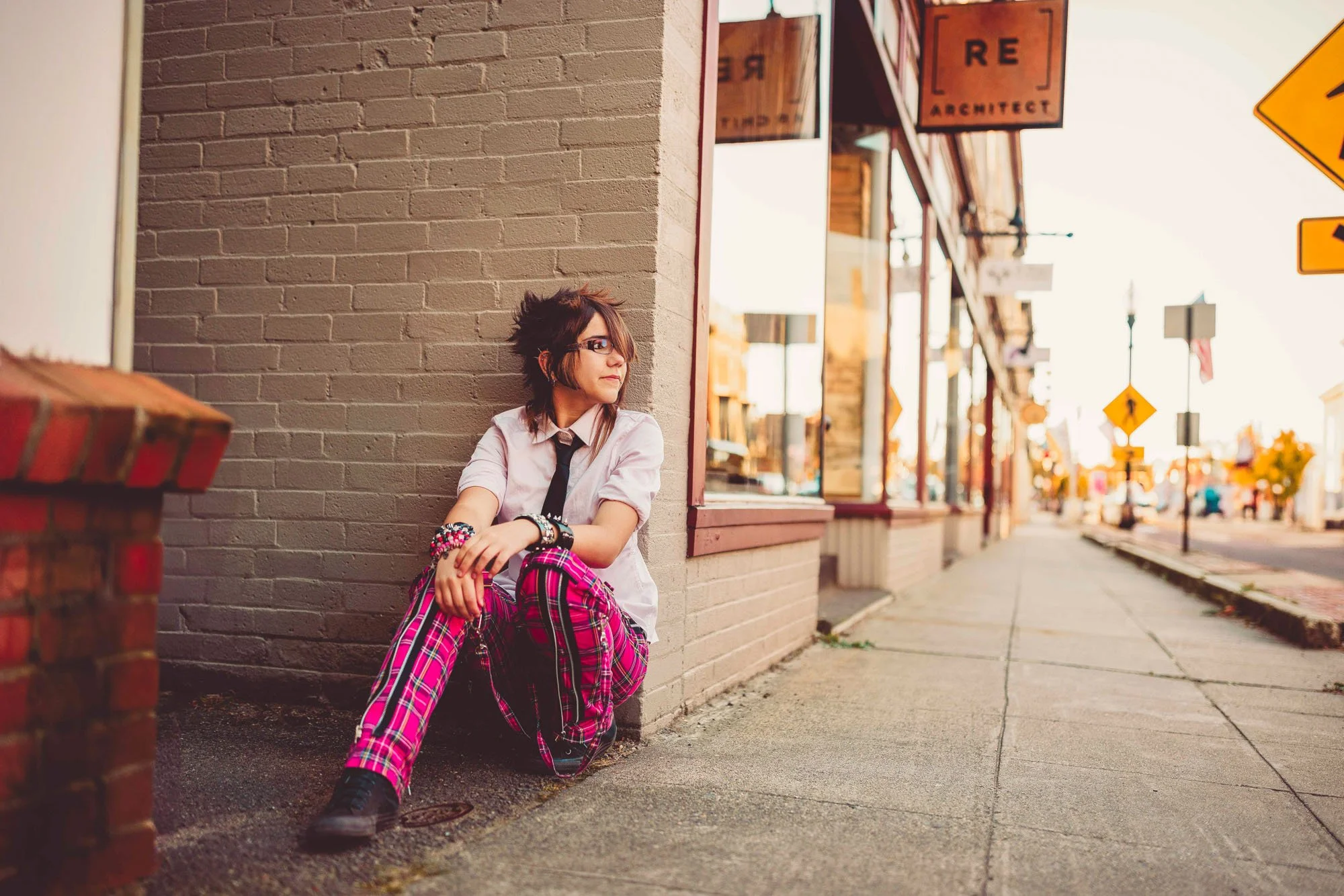 Young woman with glasses, wearing a school uniform and pink plaid pants, sitting on the sidewalk against a brick wall in a small town or city street as the sun sets.