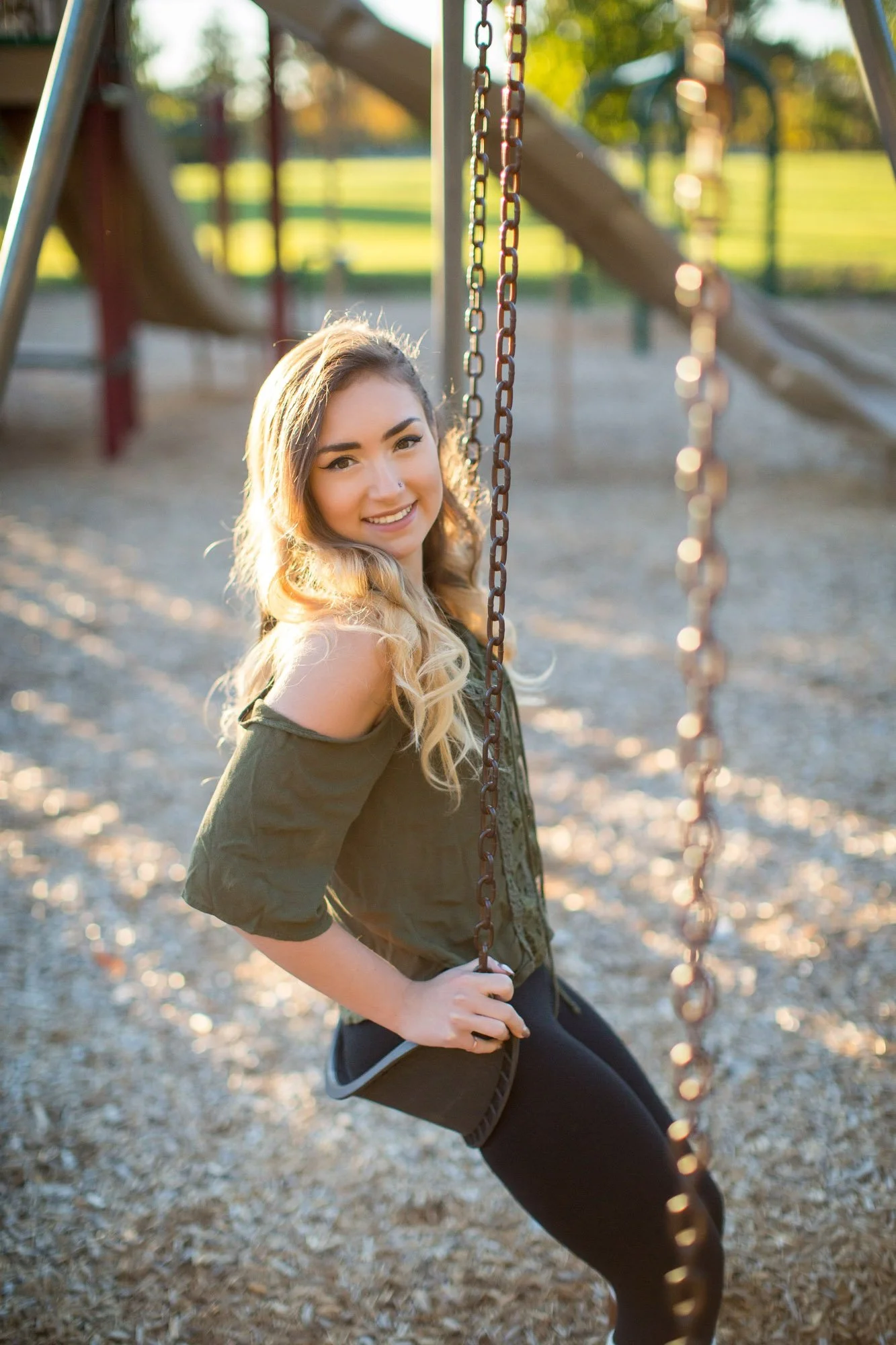 A young woman with long, wavy blonde hair and a smile, sitting on a swing at a park during sunset.