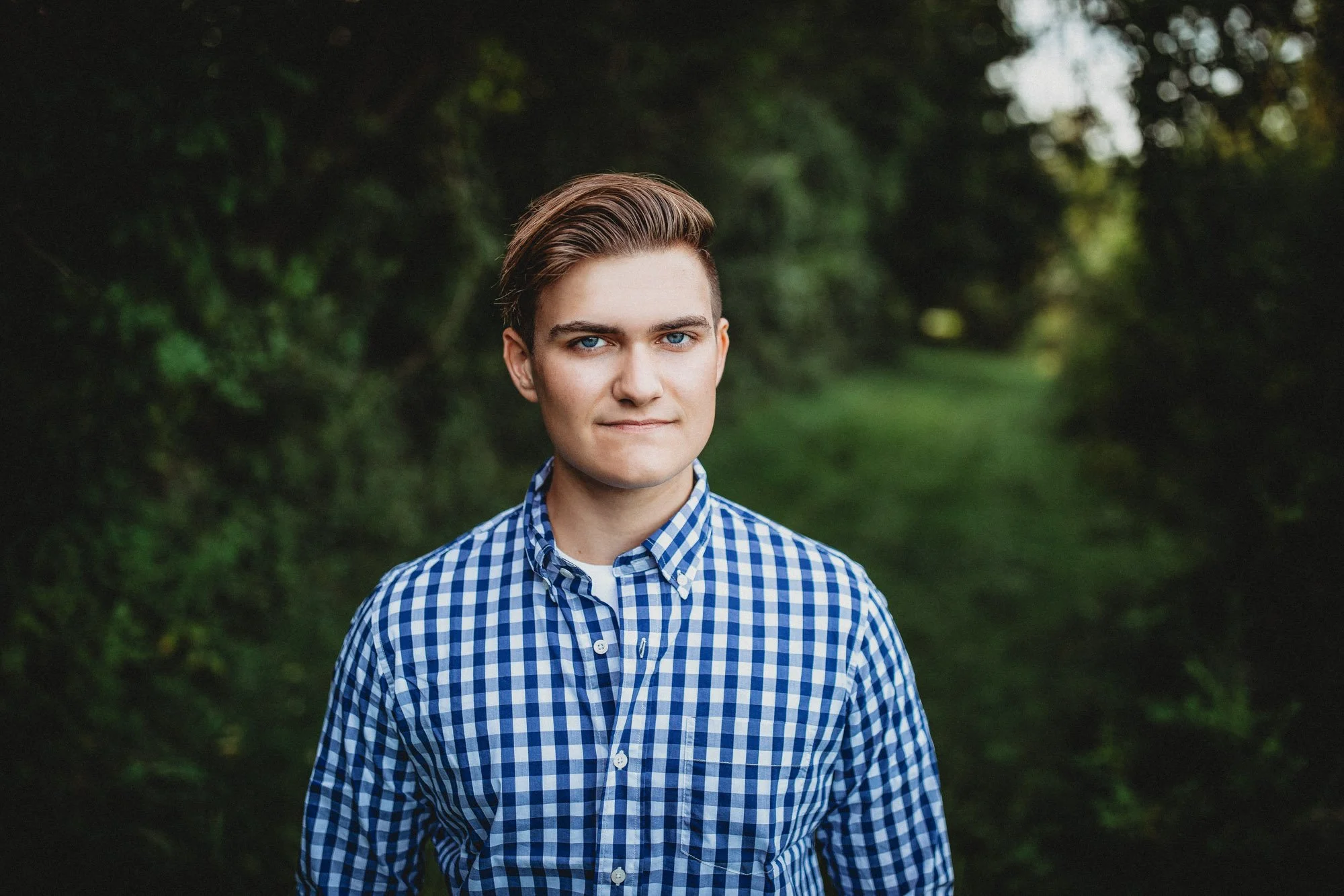 A young man with light skin, brown hair, and blue eyes standing outdoors in a green, wooded area, wearing a blue and white checkered shirt.