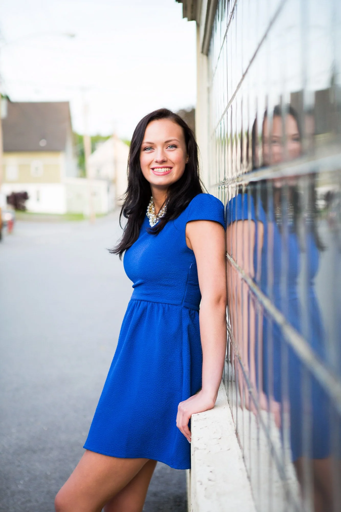 A young woman with black hair in a blue dress standing on a sidewalk, smiling, with her back against a tiled wall, and her reflection visible in the tiles.
