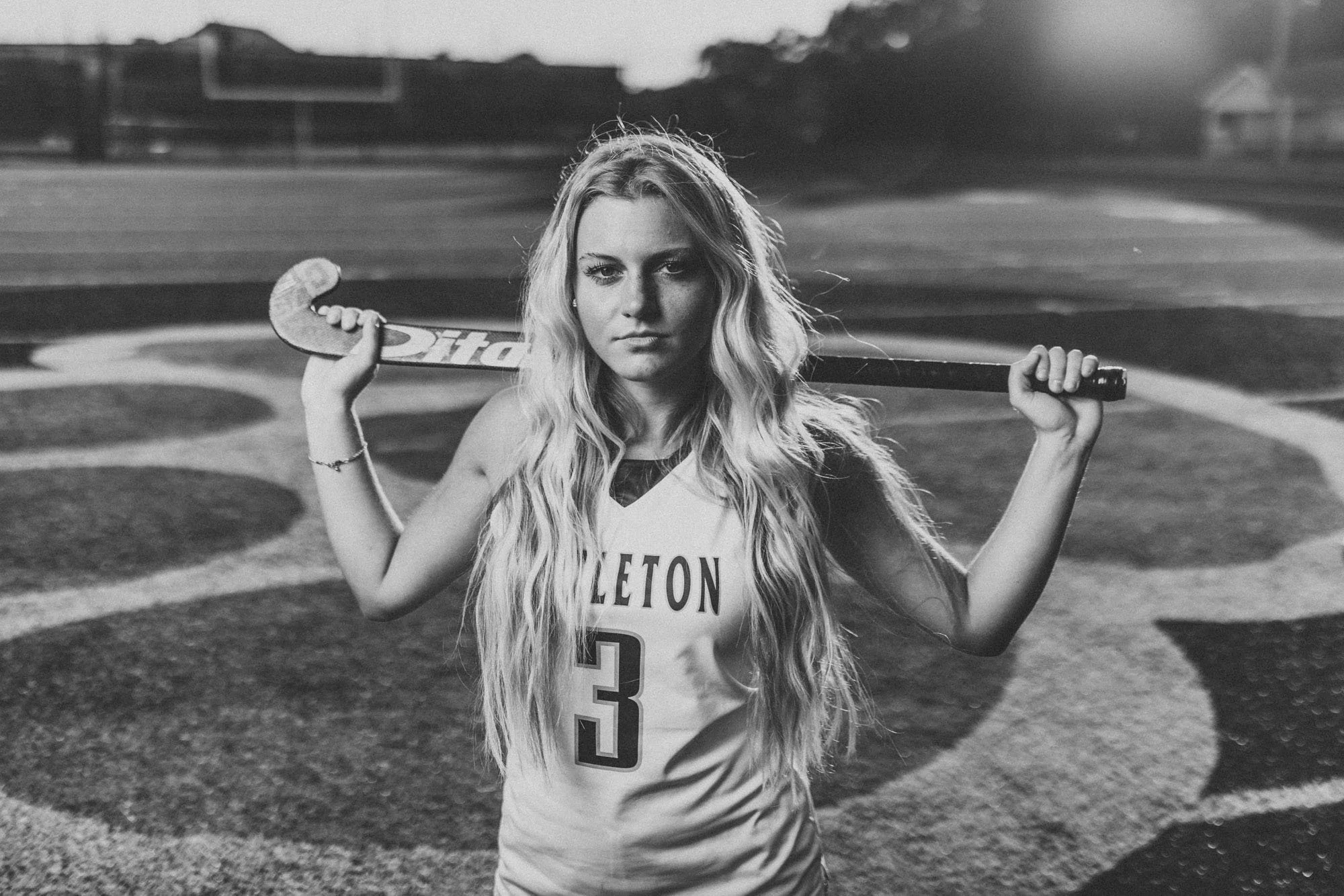 A young woman in a sports jersey holding a hockey stick across her shoulders, standing on a sports field during sunset.