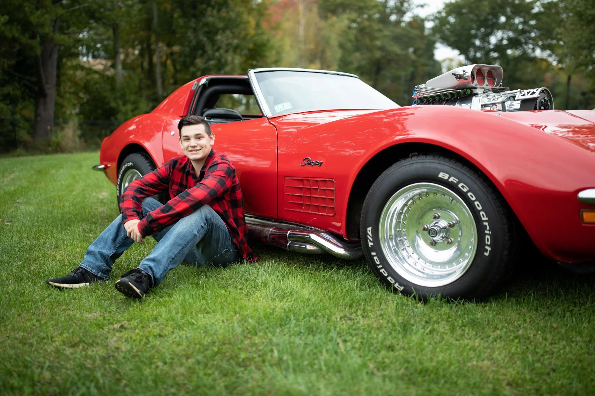 A young man in a red and black plaid shirt sitting on green grass next to a red sports car in an outdoor setting with trees in the background.