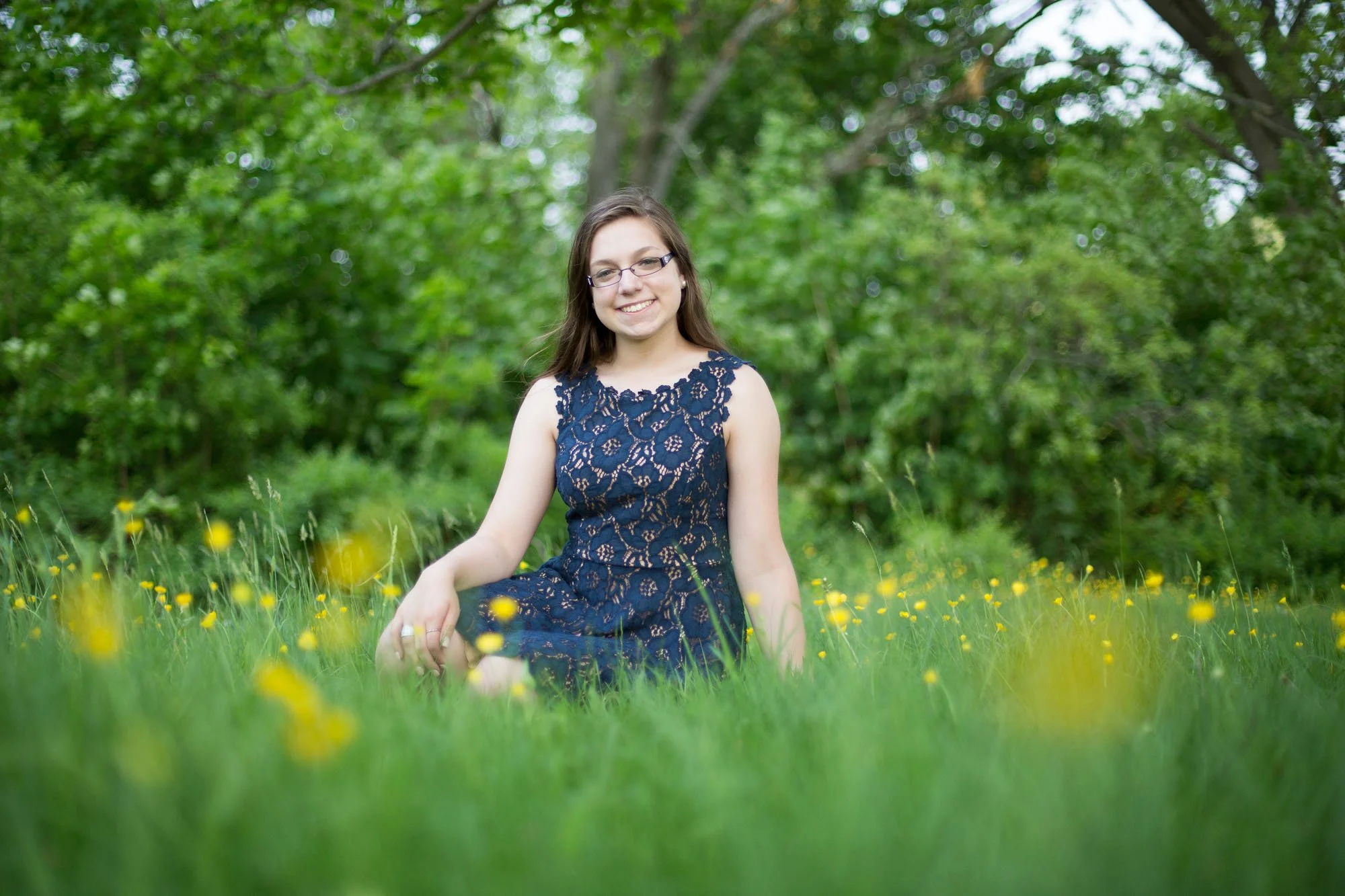 Smiling woman in a navy lace dress sitting in a grassy field with yellow flowers, surrounded by green trees.