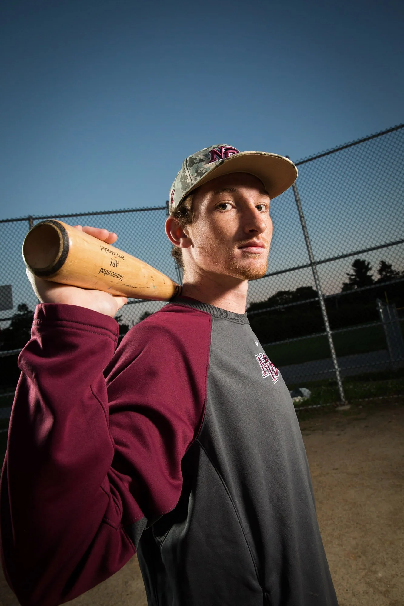 A young man holding a baseball bat over his shoulder at a baseball field during dusk, wearing a cap and athletic shirt.