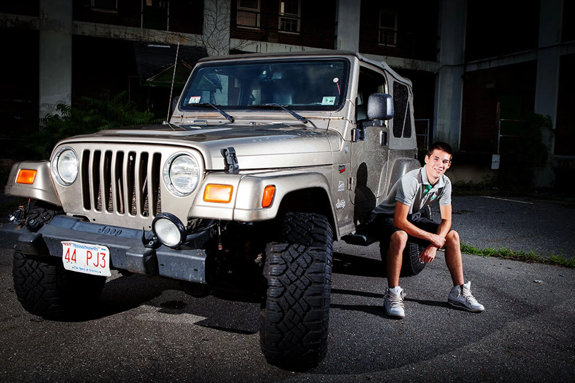 A young man posing next to a beige Jeep Wrangler with Massachusetts license plates, parked on a dark asphalt surface at night, with a building in the background.