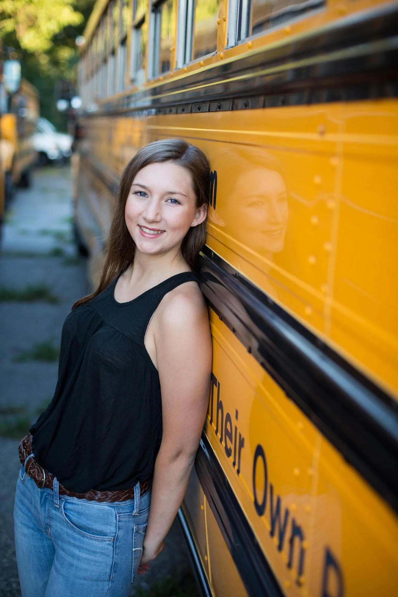 A young woman with long brown hair, wearing a black sleeveless top and blue jeans, leaning against a yellow school bus and smiling at the camera.